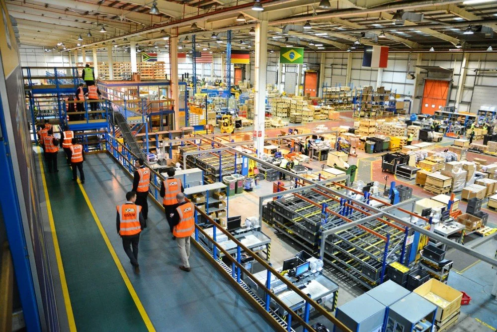 Inside a large warehouse with workers wearing orange safety vests. The warehouse contains shelves filled with boxes and equipment, and flags from various countries hanging from the ceiling.