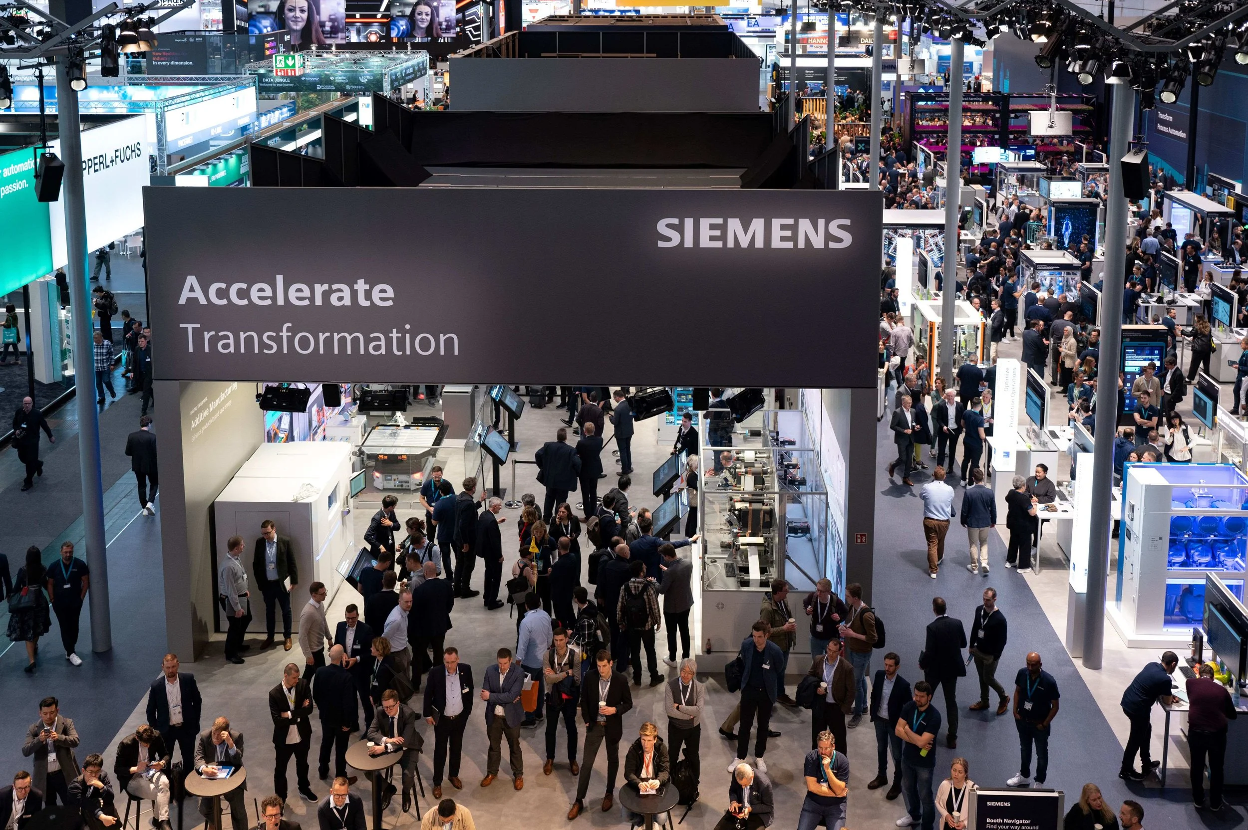 A crowded trade show or exhibition with Siemens booth displaying the tagline 'Accelerate Transformation'. Numerous attendees are walking, talking, and exploring booths and displays.
