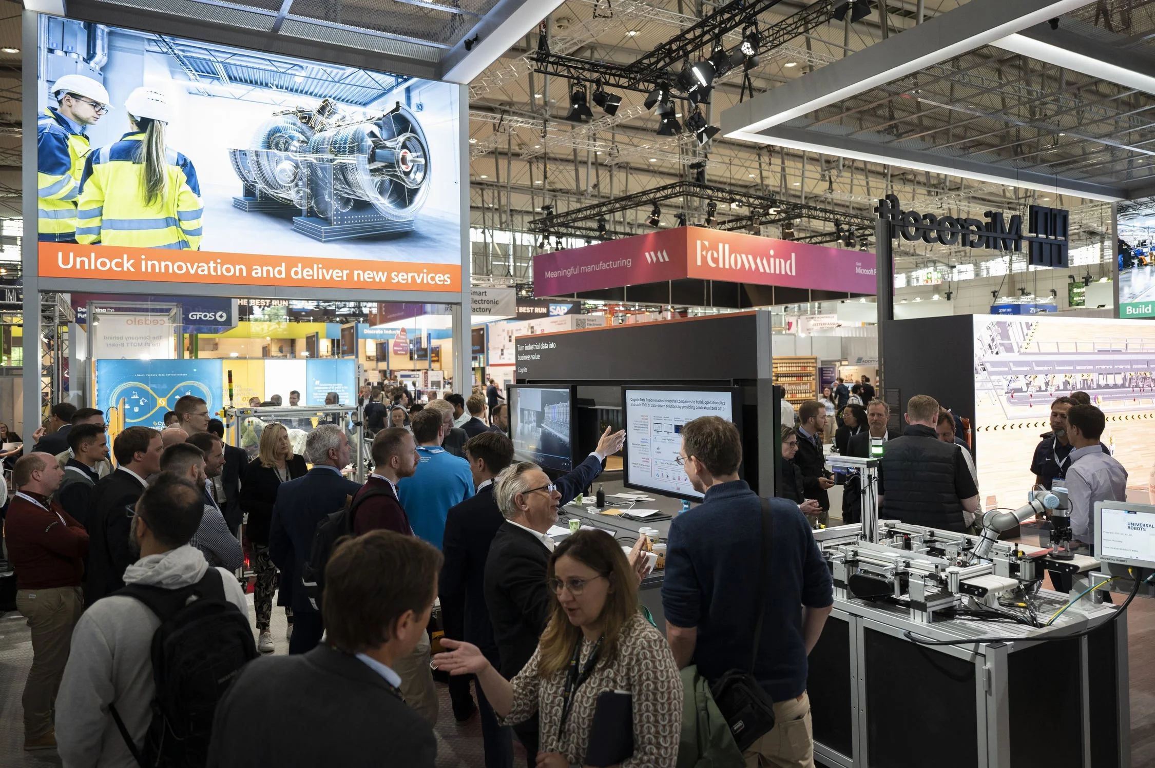 A crowded trade show with various booths and displays, including a large screen and machinery, with people engaging in discussions and presentations.