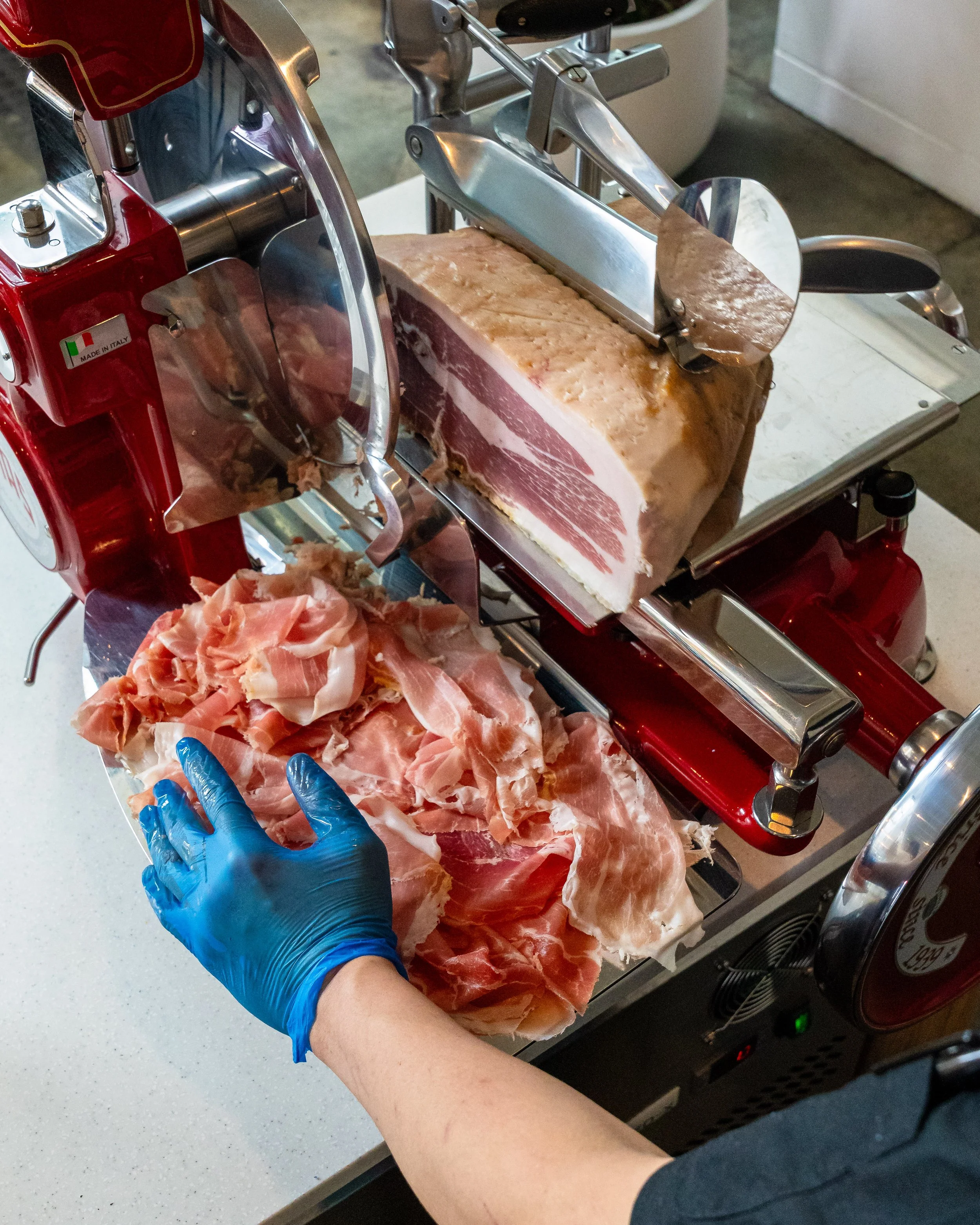 A person in a blue glove slicing cured ham on a meat cutter, with slices of ham on a tray nearby.