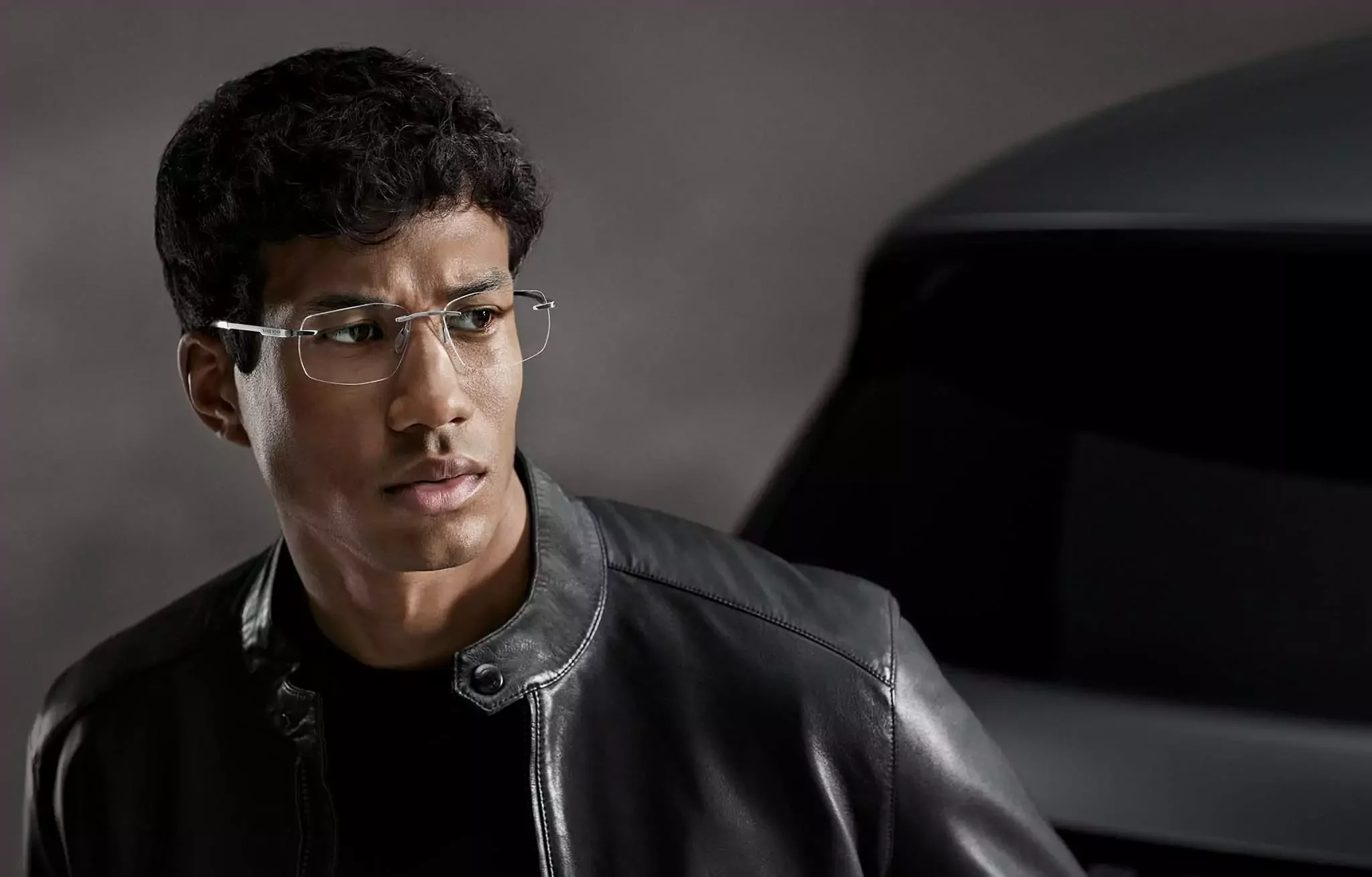 A young man with glasses and curly hair looks to his left, wearing a black leather jacket, with a dark background and a blurred object beside him.
