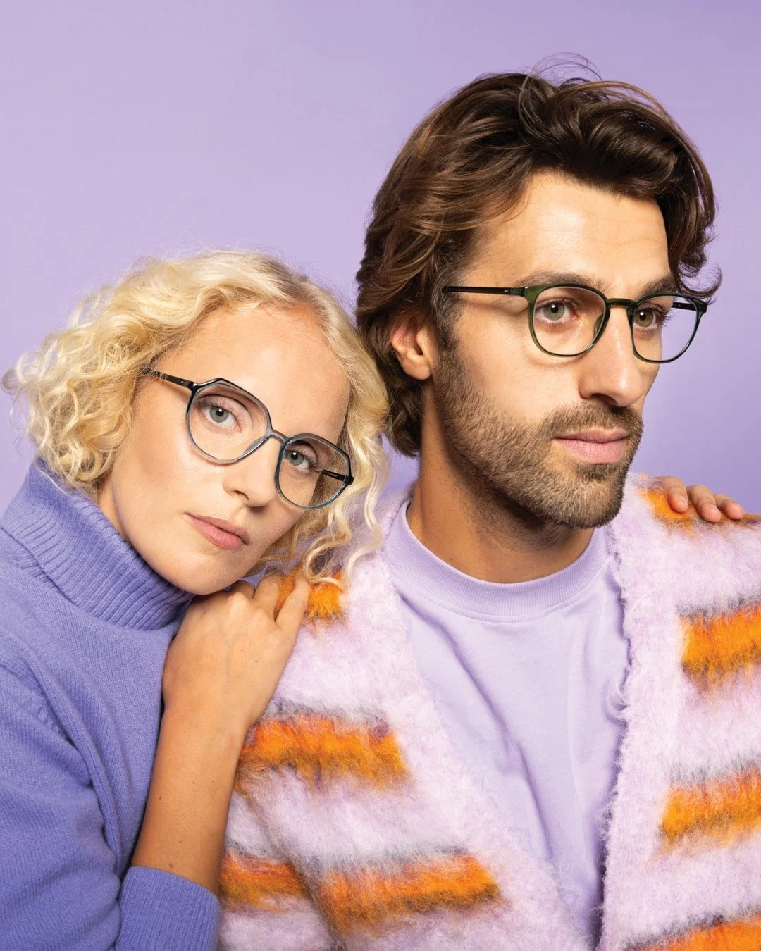 A woman with curly blond hair and glasses, wearing a purple turtleneck, leaning on a man with dark hair and glasses, wearing a fuzzy striped sweater with purple, orange, and white colors, against a purple background.
