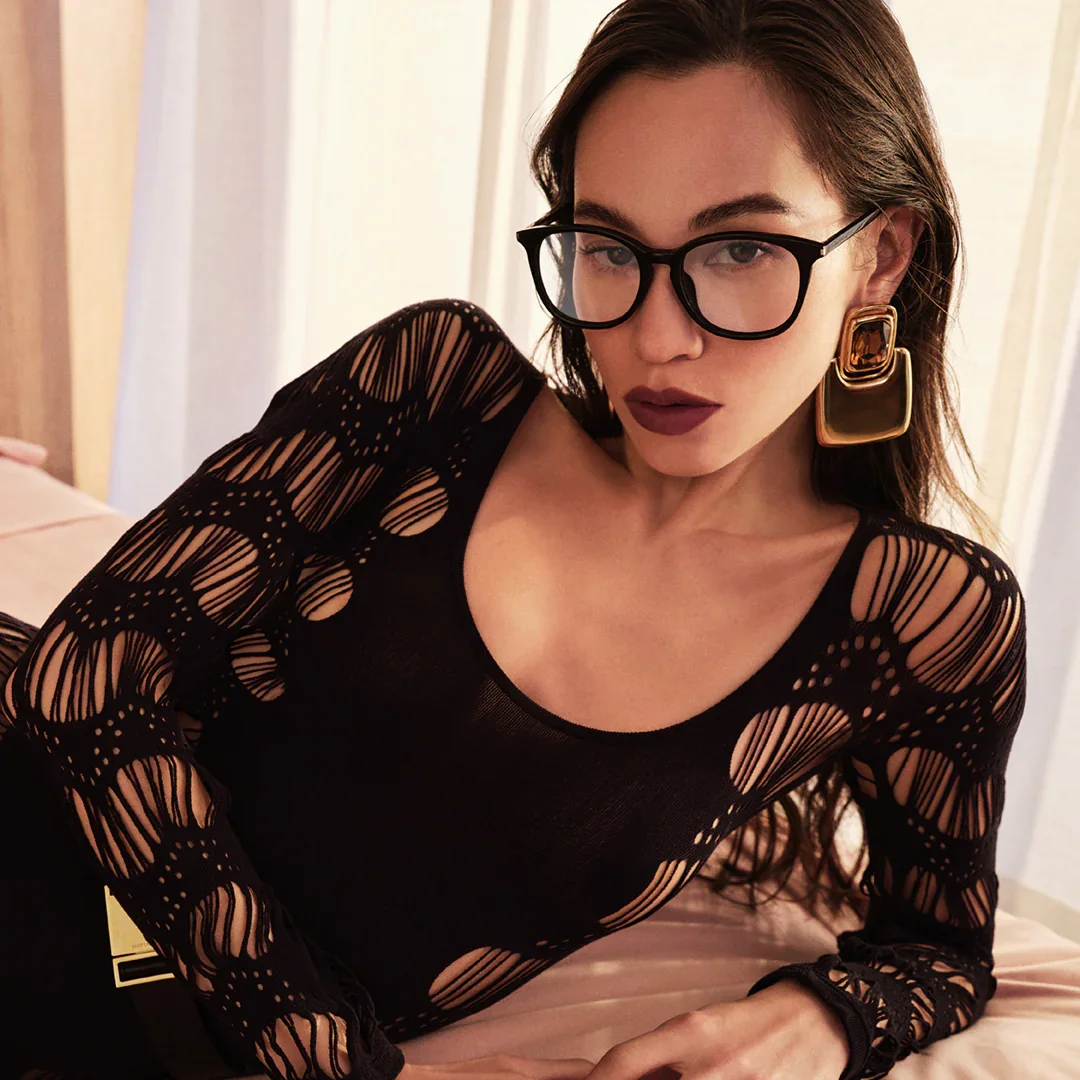 Woman with dark brown hair wearing large black glasses, statement gold earrings, and a black lace dress with intricate cutout patterns, lying on a bed in a softly lit room.
