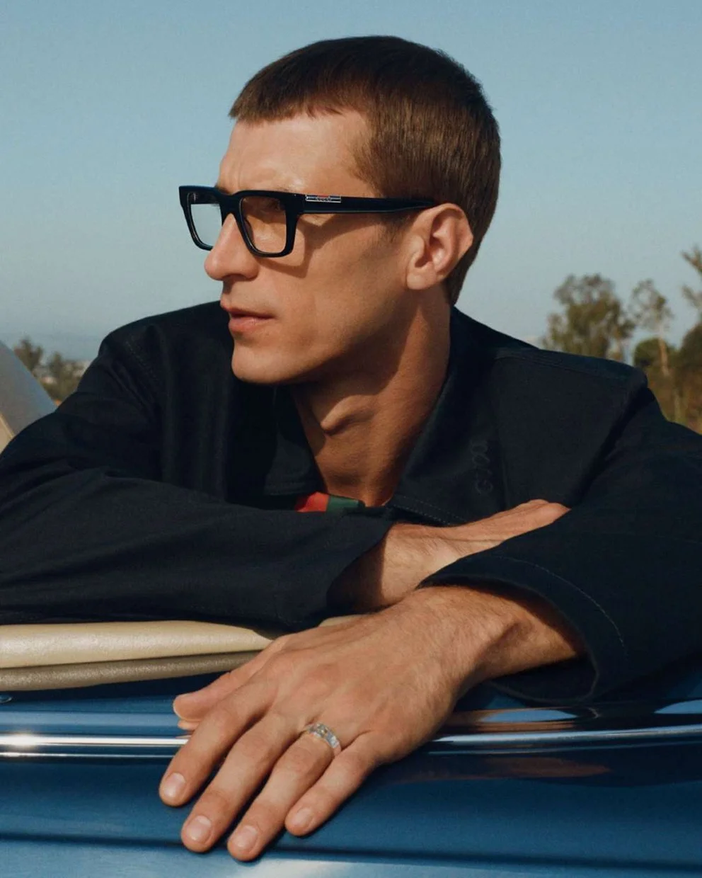 A young man with short brown hair, wearing black glasses and a black jacket, is resting his arms on a metallic surface outdoors during daytime, with a clear sky and some trees in the background.