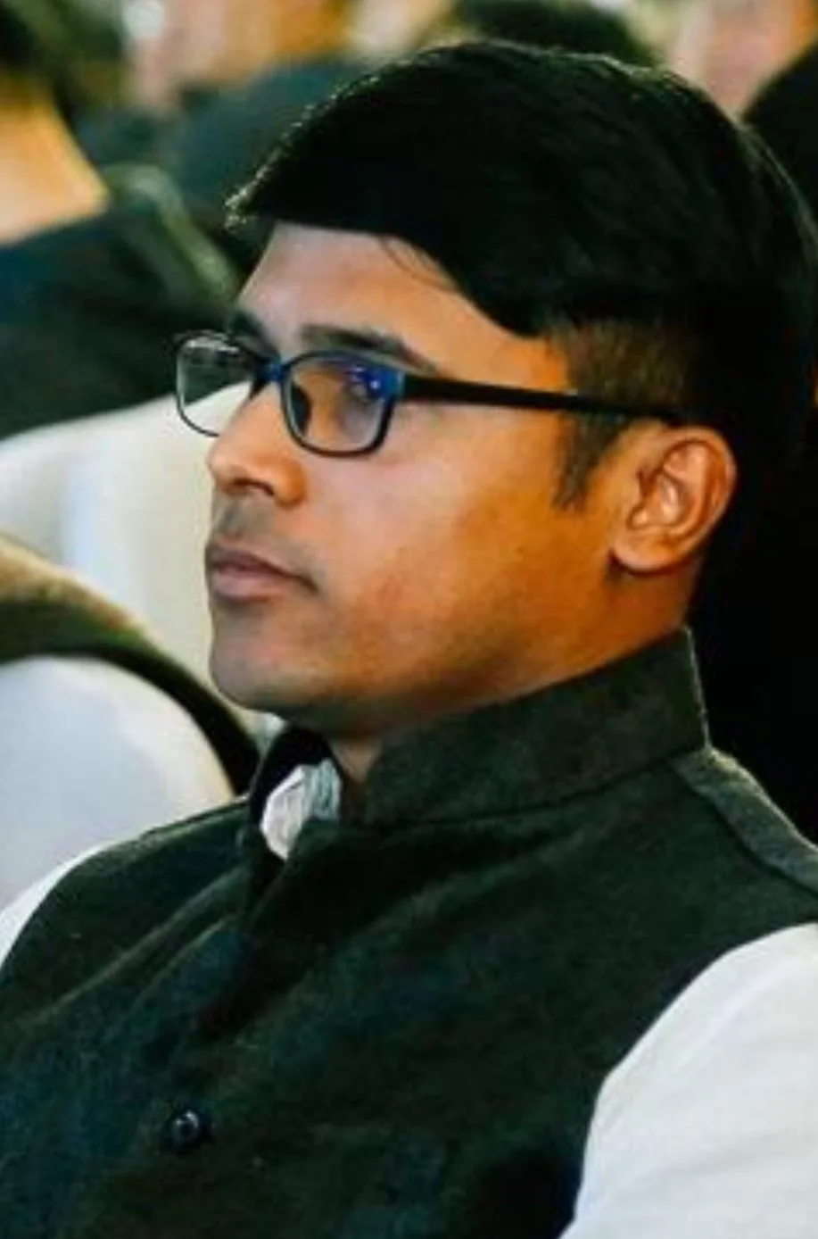 A man with black hair wearing glasses, a black vest, and a white shirt, sitting and looking to the side at an indoor event.