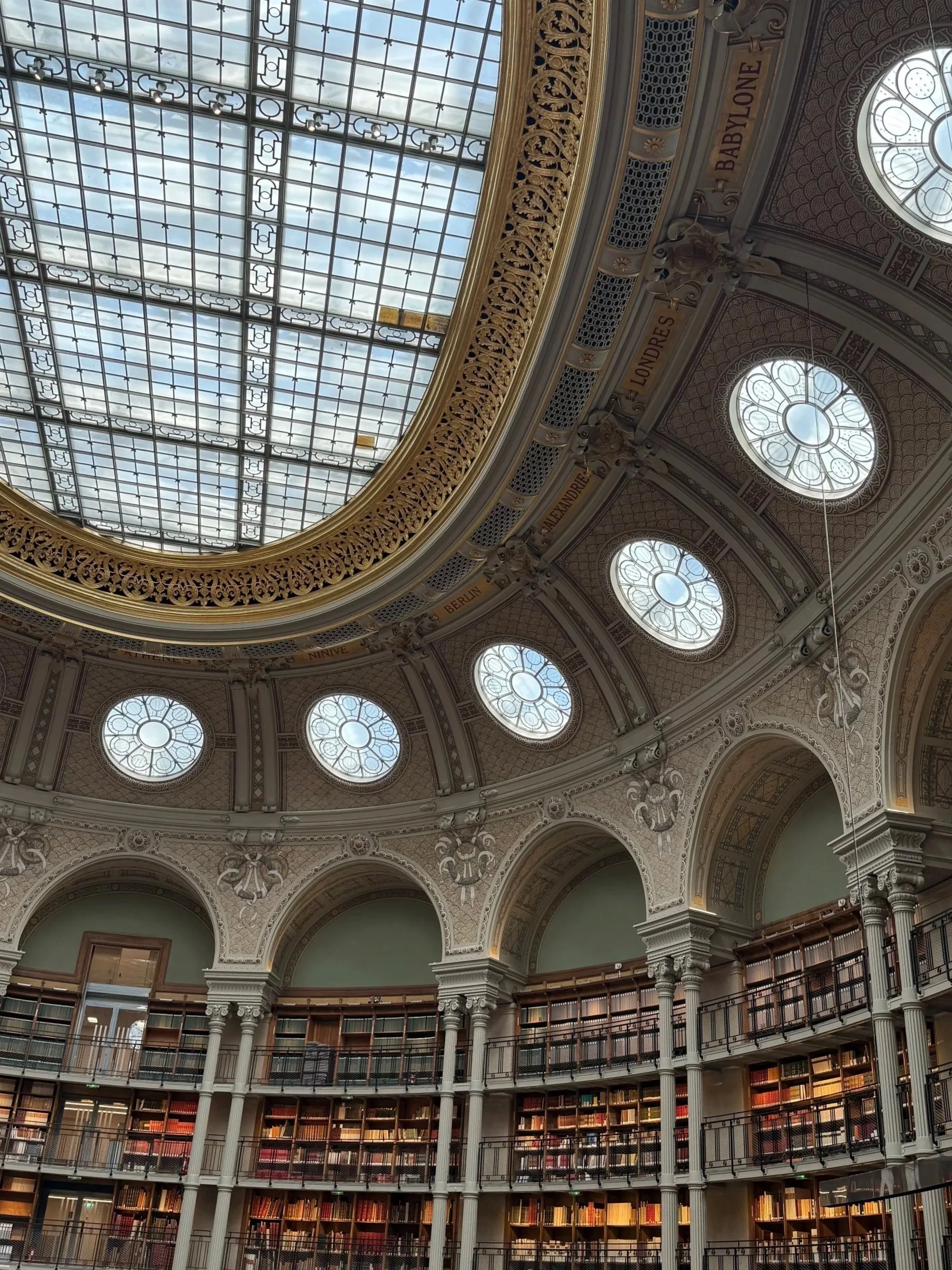 Intérieur d'une bibliothèque élégante avec un plafond en verre en forme de dôme et plusieurs niveaux de rayonnages remplis de livres.