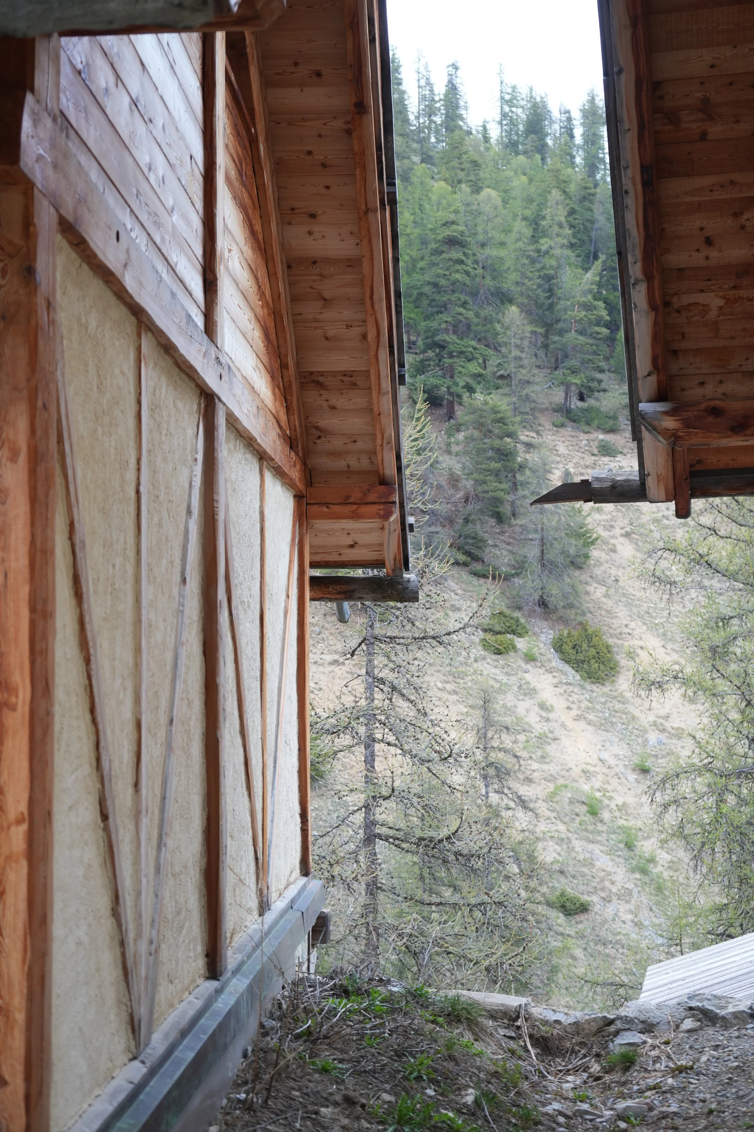 Vue extérieure d'une construction en bois, avec un mur partiellement isolé et un cadre de fenêtre sans vitre, donnant sur une forêt montagneuse.
