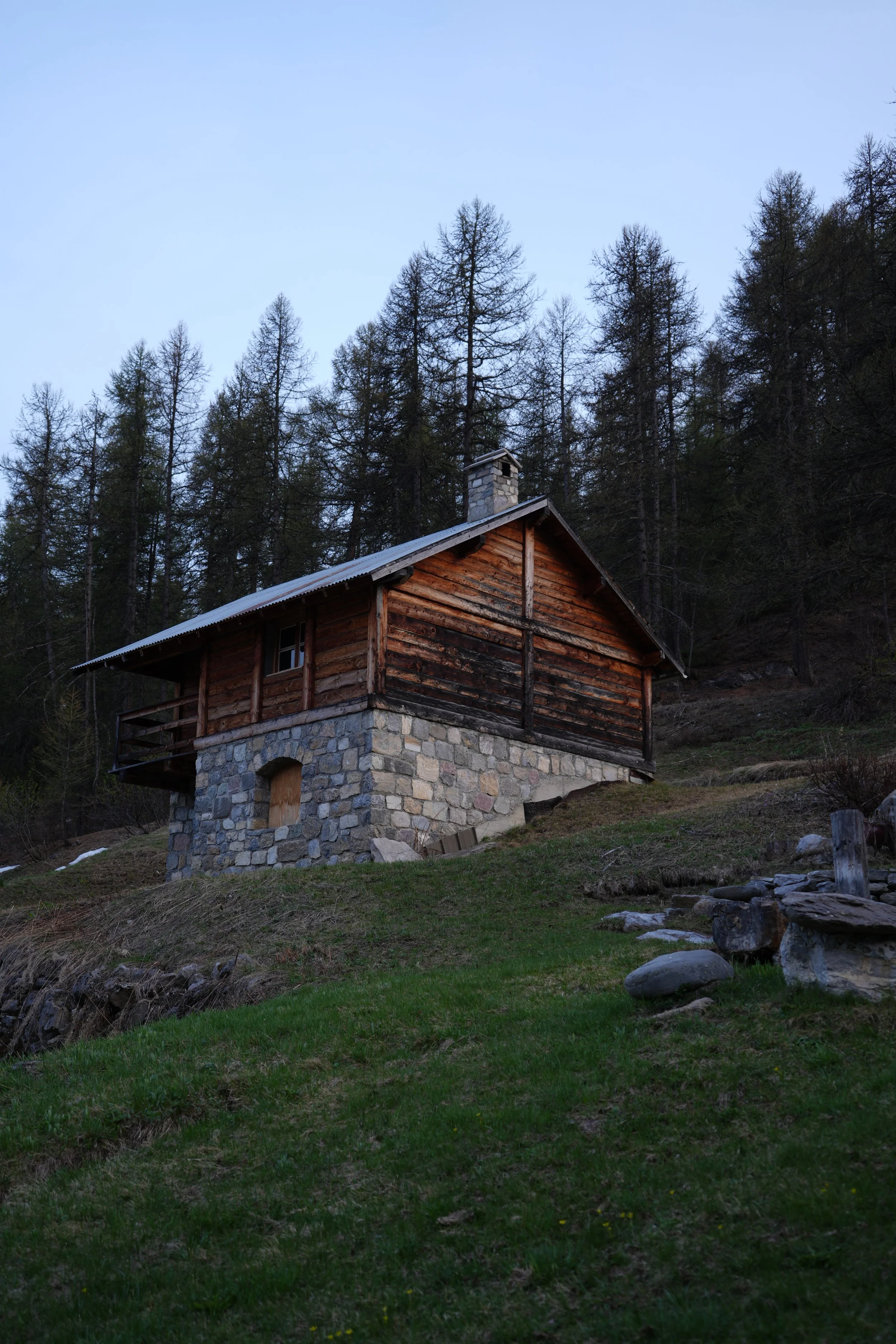 Cabane en bois avec une fondation en pierre, située sur une pente herbeuse entourée d'arbres, sous un ciel clair.