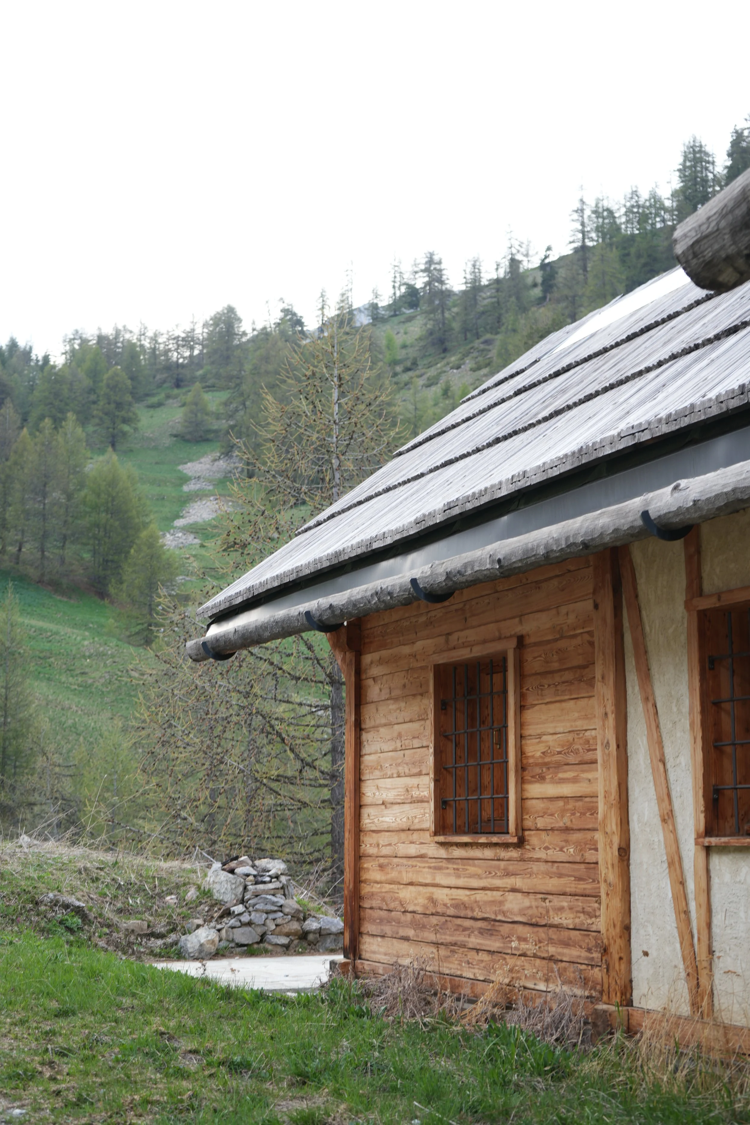 Une maison en bois avec un toit en tuiles et un mur en pierre, située dans un paysage montagnard verdoyant avec des arbres et une pente escarpée en arrière-plan.