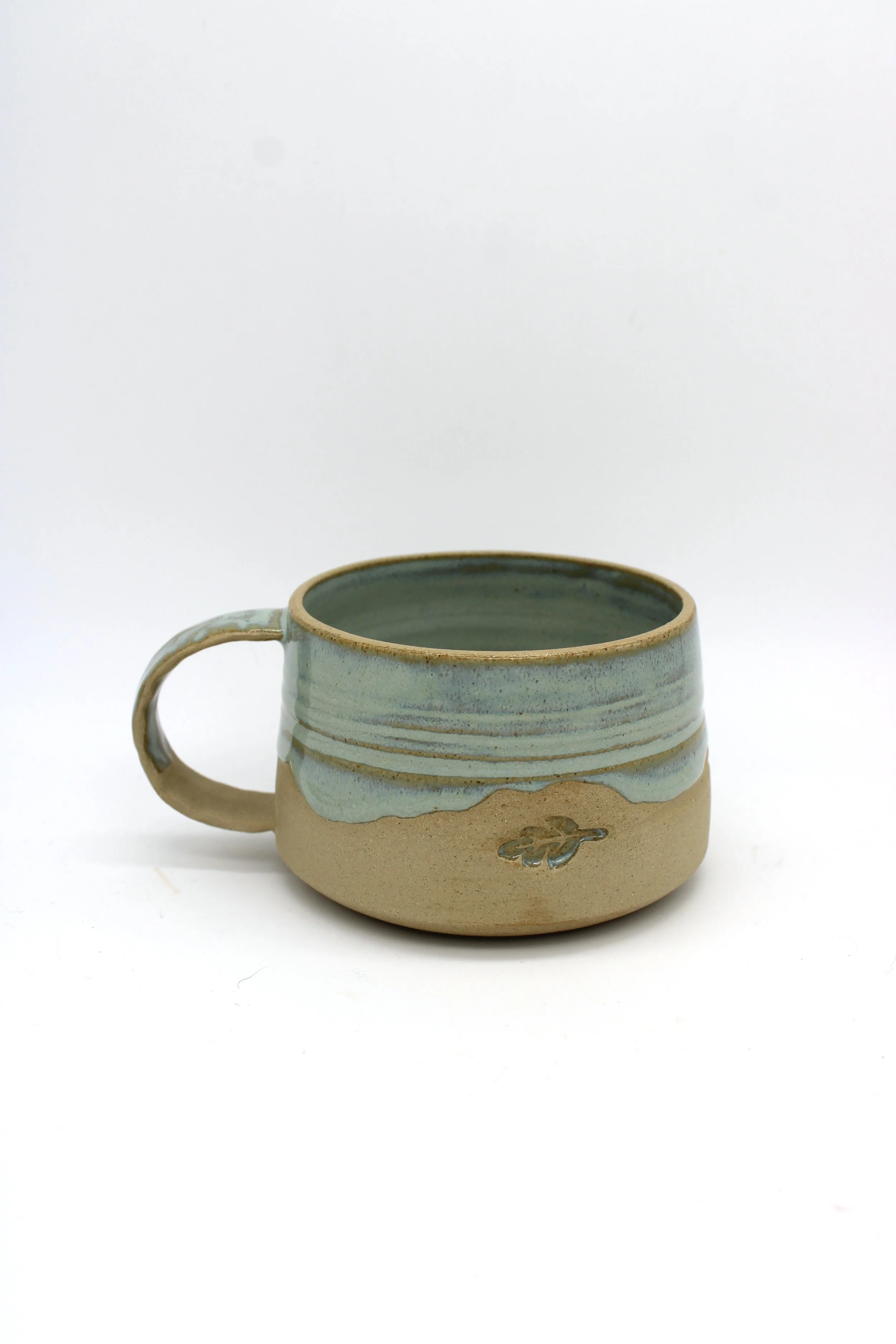 A ceramic coffee mug with blue and sandy beige glaze and a small turtle design on the front, placed on a white background.