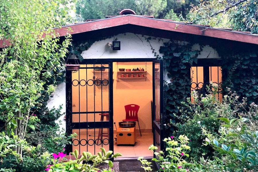 A small house with a red roof, white walls, and black window frames. The front door is open, revealing a chair and a shelf with pottery inside. The house is surrounded by lush greenery and flowering plants.