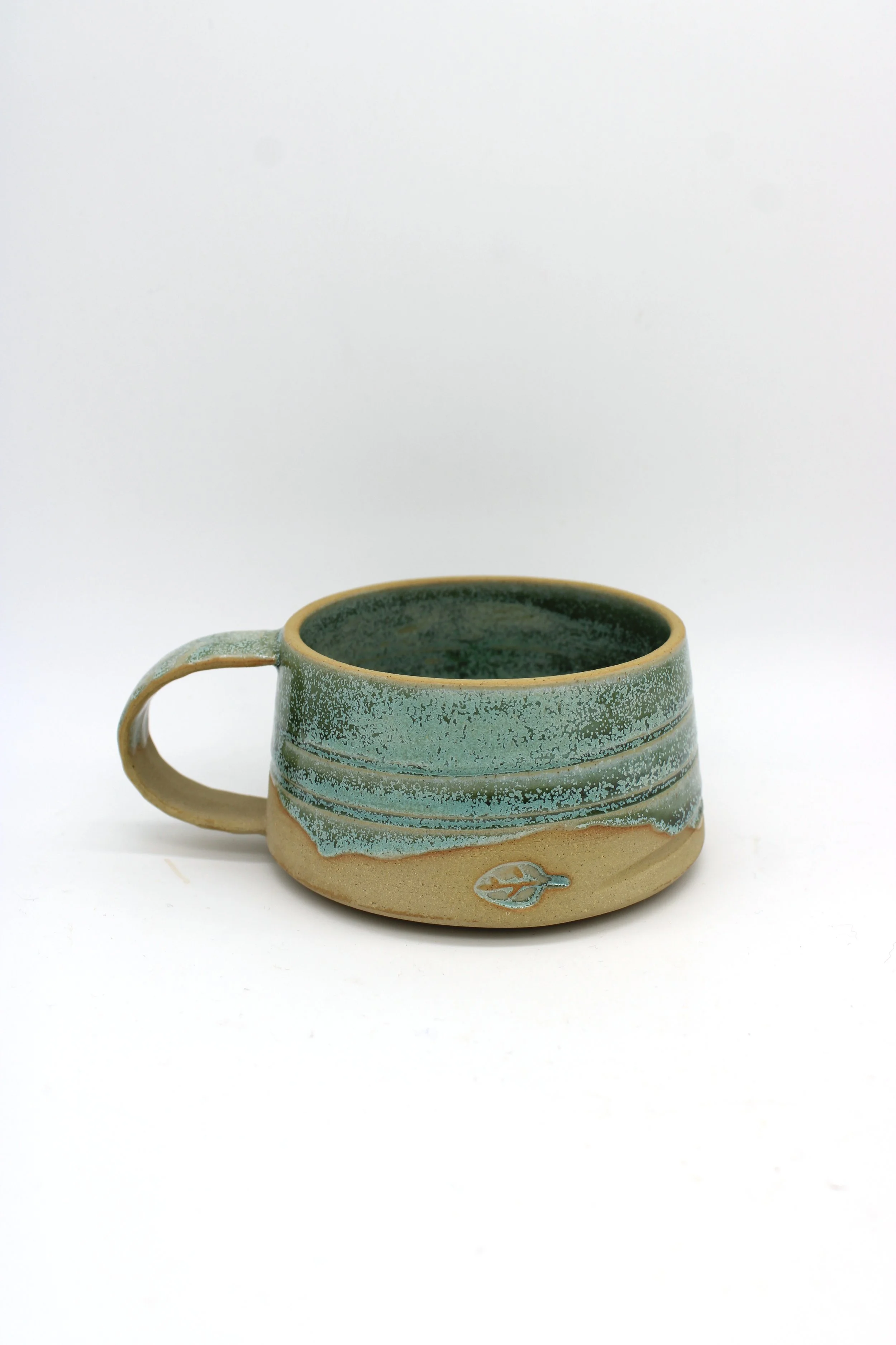 Ceramic mug with a natural beige base, decorated with teal glaze and a leaf emblem on the side, placed on a white background.
