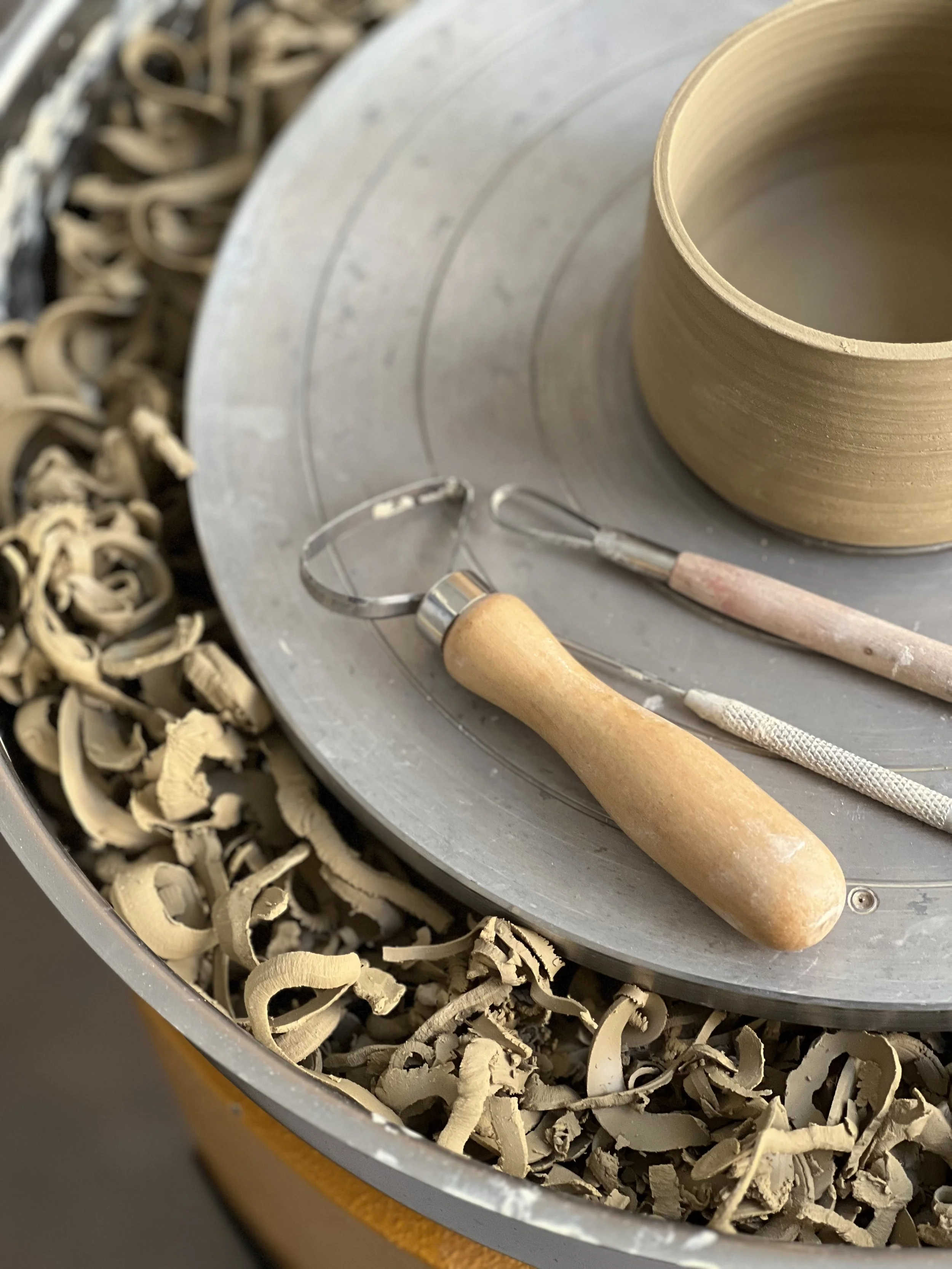 Close-up of a pottery wheel with a partially shaped clay pot, pottery tools, and wood shavings around the wheel.