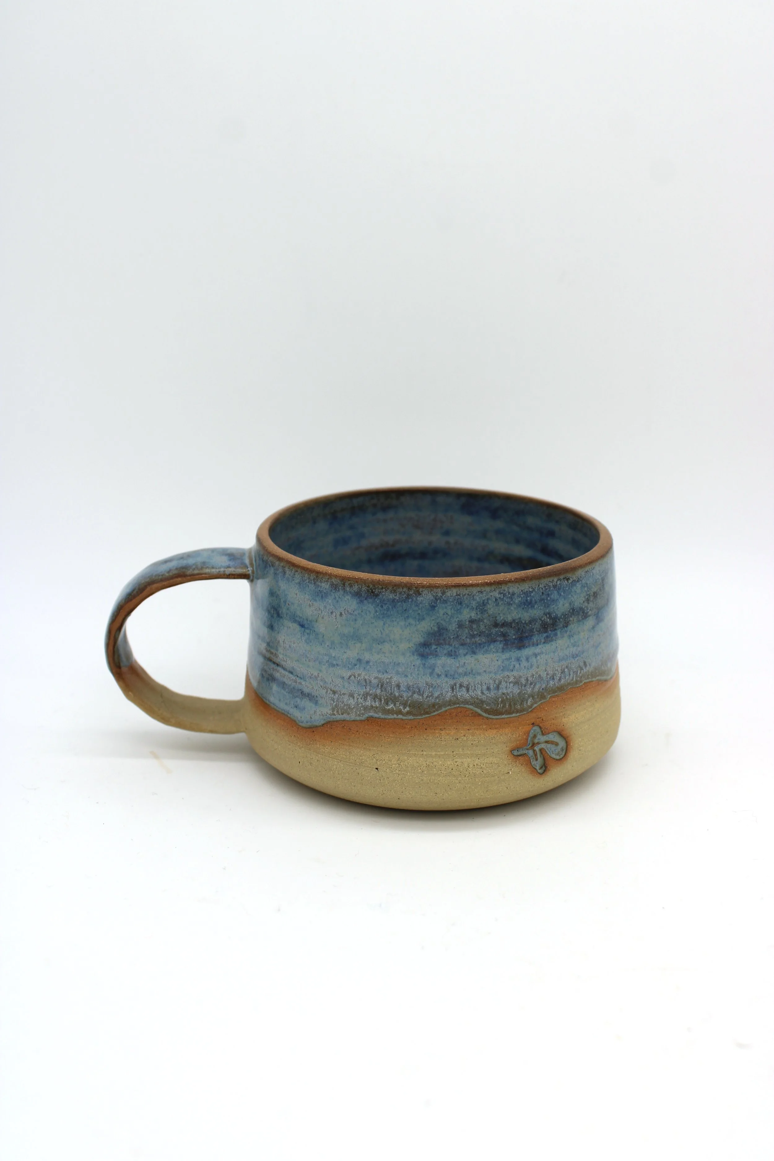 Handmade ceramic mug with a blue and beige glaze, featuring a small fish-shaped decoration on the side, placed against a plain white background.