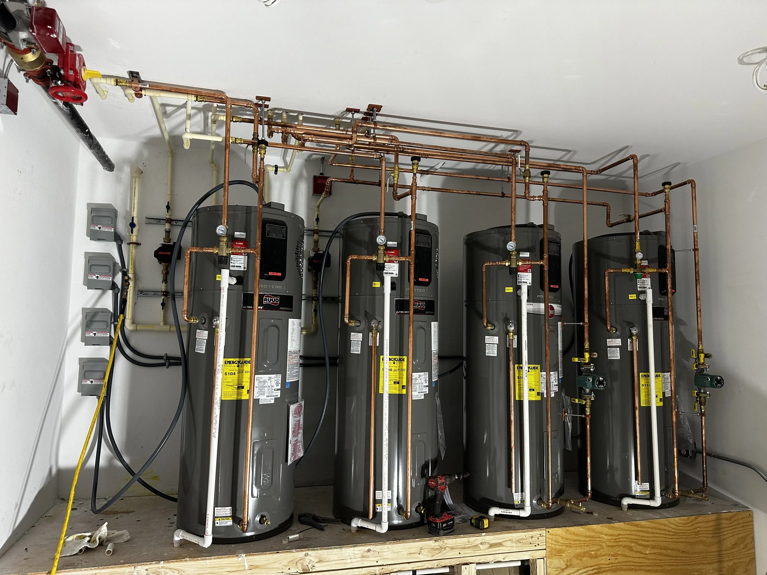 A row of five water heater tanks with piping and valves, placed on a wooden platform in a utility room.