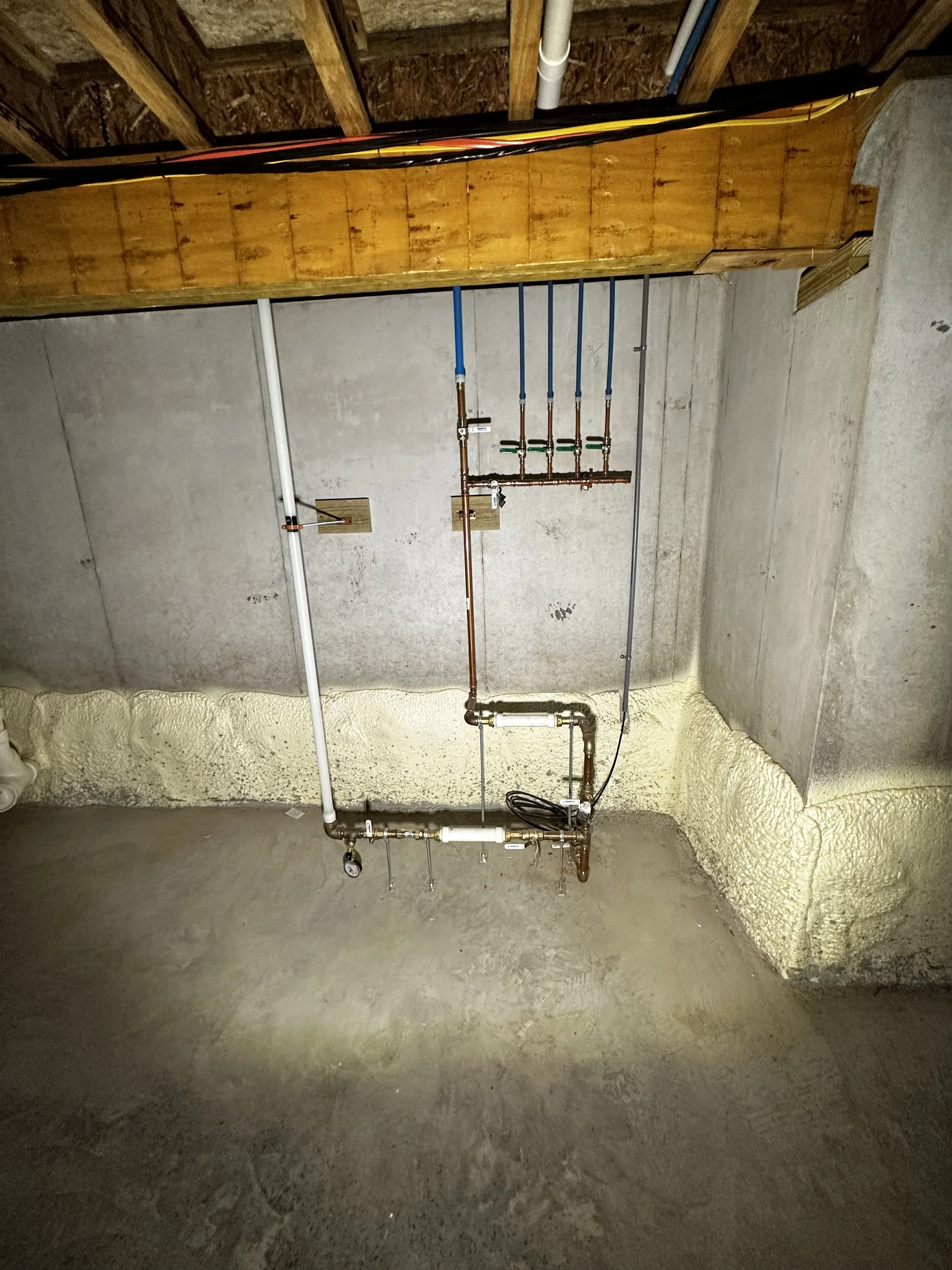 Underground plumbing pipes and fittings in a basement with concrete walls and floor.