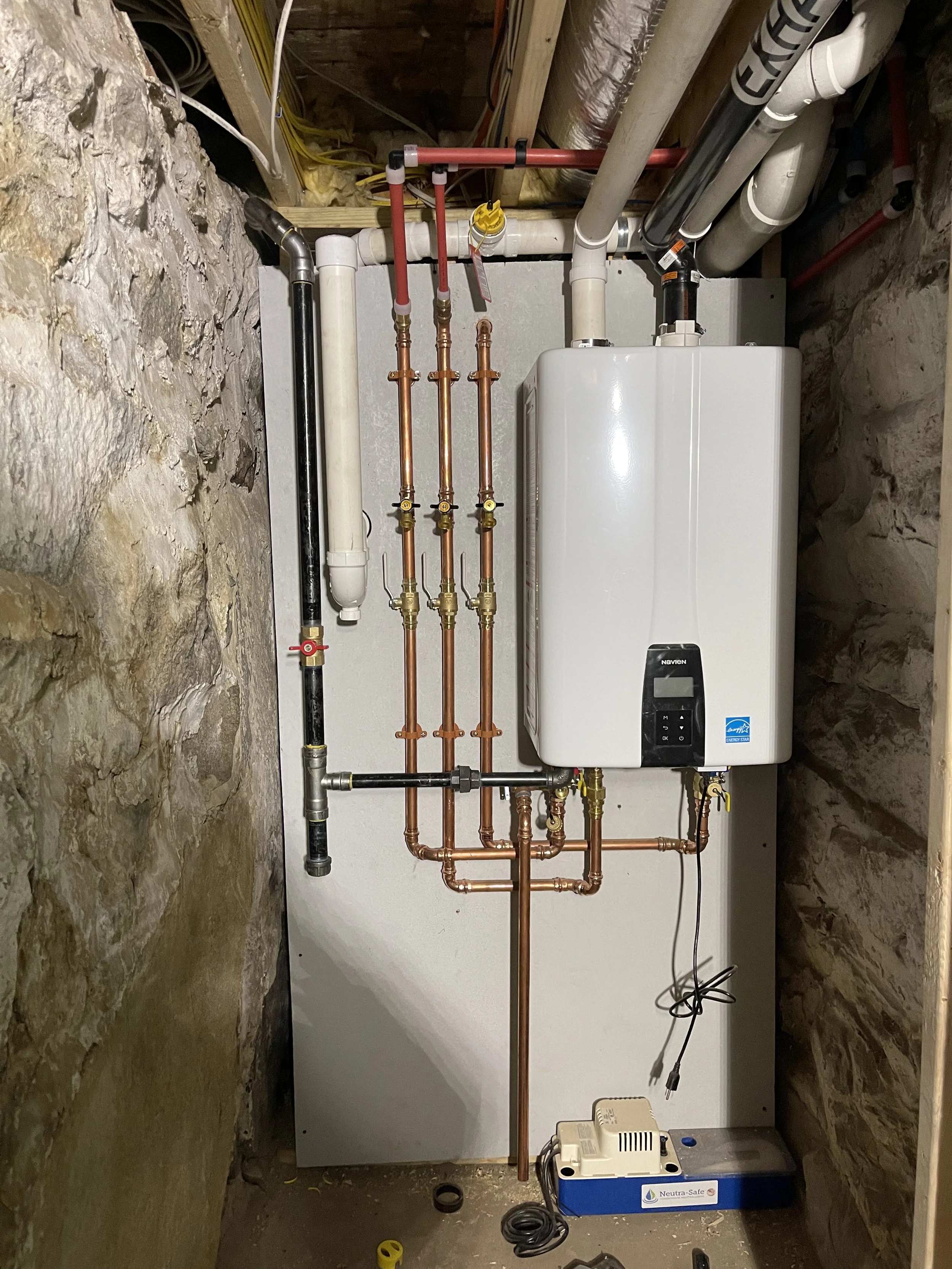 A utility room with a tankless water heater, copper and plastic piping, and various plumbing components mounted on a white wall.