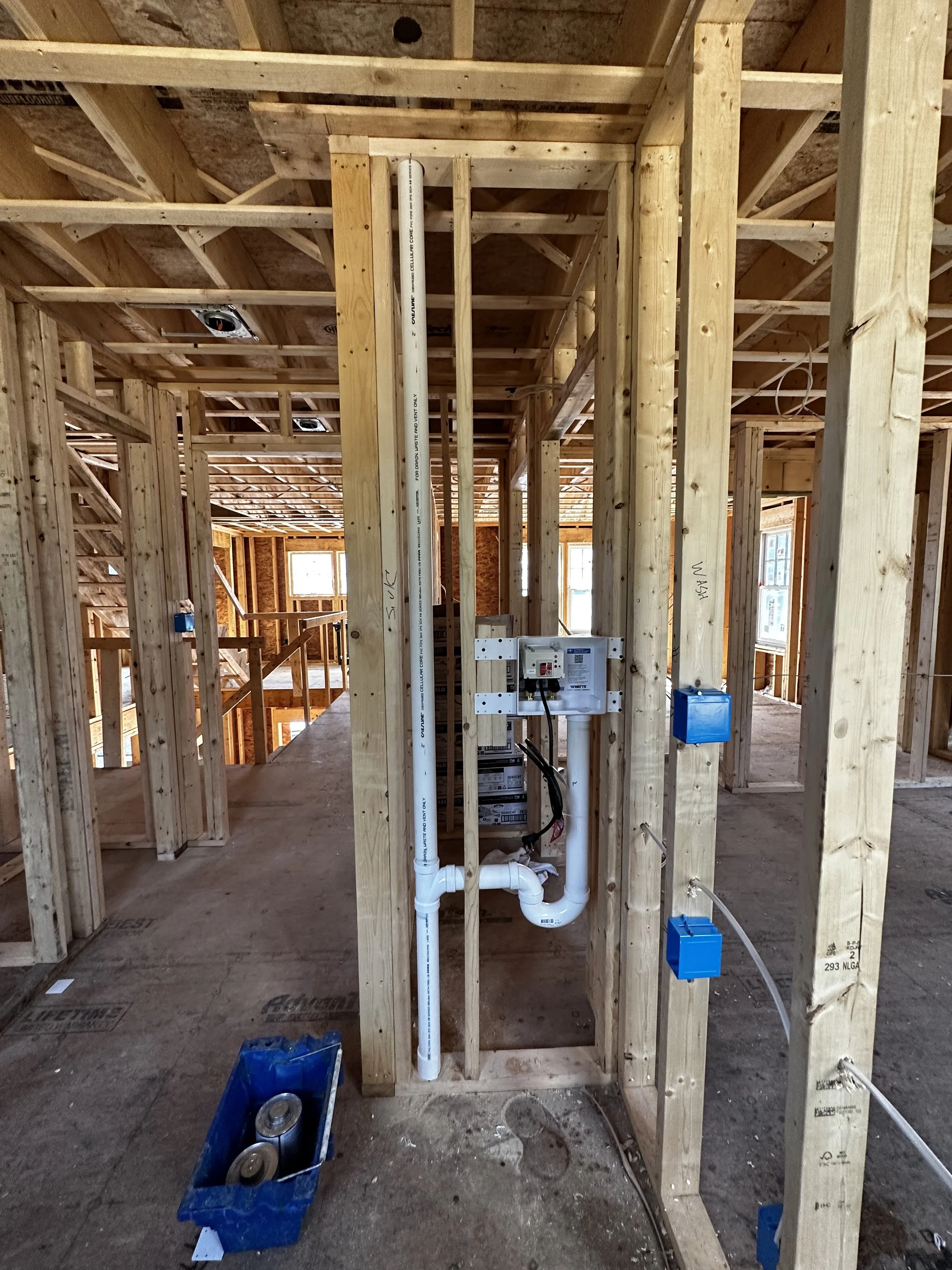 Interior of a building under construction with exposed wooden framing, electrical wiring, and plumbing pipes in the wall. No finished walls or flooring are visible.