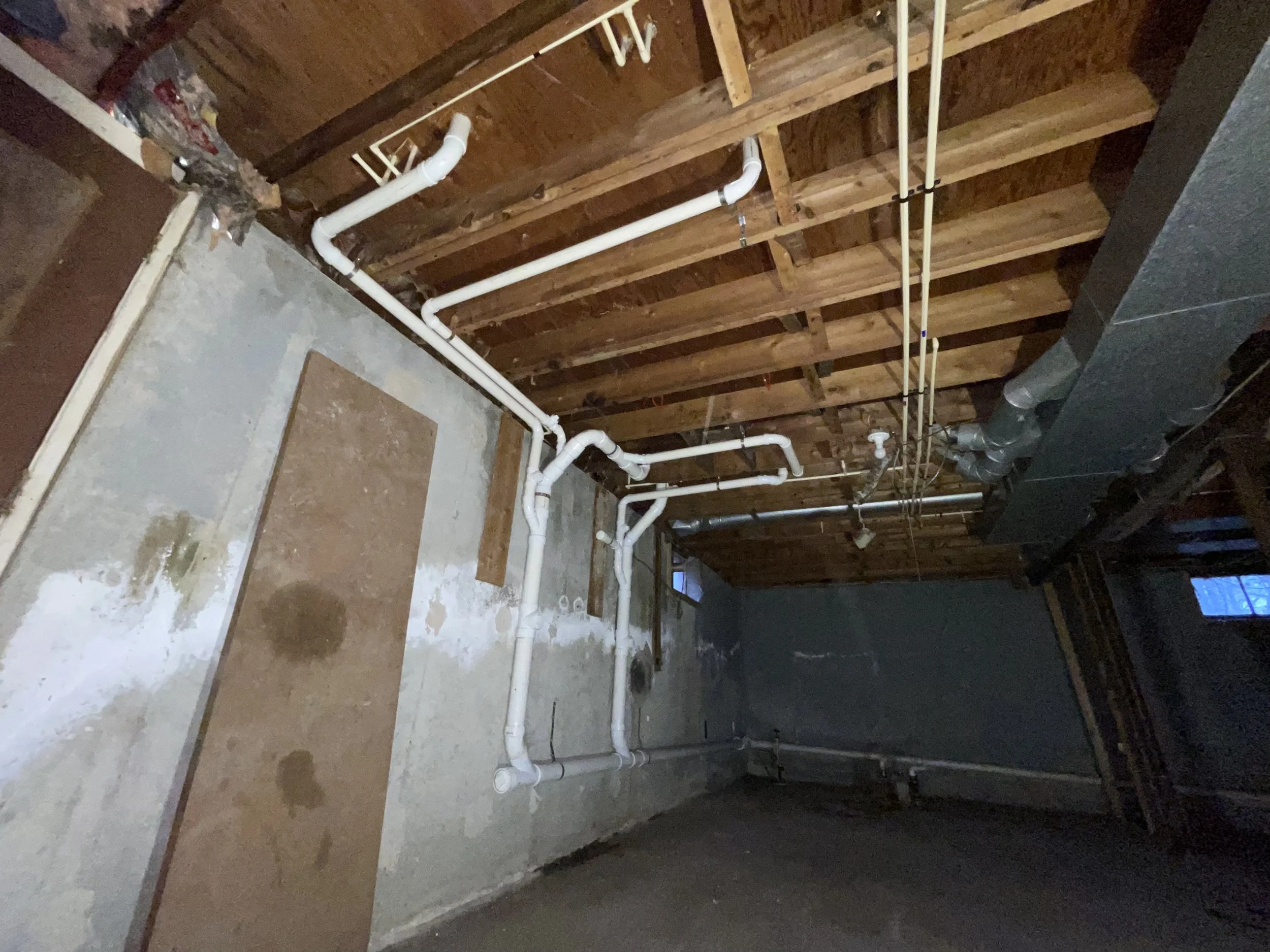 Construction site showing exposed wooden ceiling beams, gray concrete wall, and white plumbing pipes installed along the wall and ceiling.