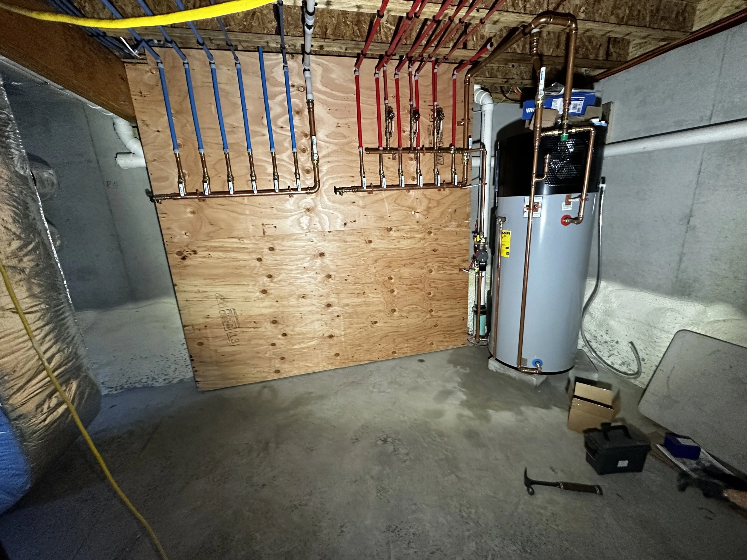 A basement with a wooden panel and plumbing pipes, a water heater, and construction tools.