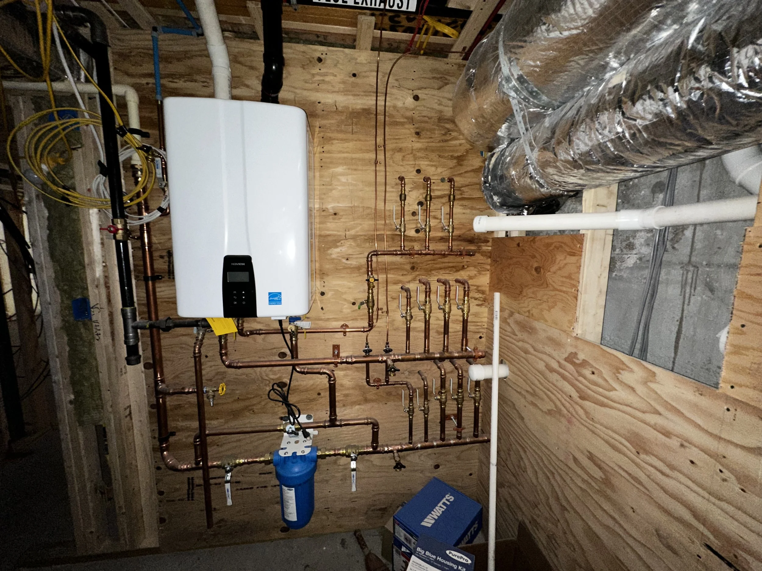 Indoor view of a plumbing system with copper pipes, a water heater, and filters mounted on a wooden wall in a utility room or basement.