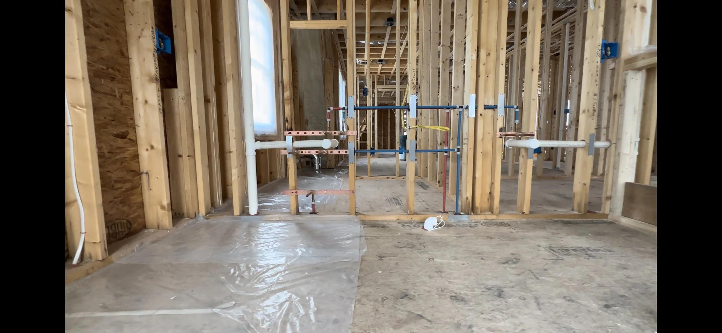 Construction site with wooden framing and plumbing pipes, partially completed interior walls, and a clear plastic sheet covering part of the floor.