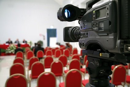 A professional video camera pointing at a conference room with red chairs and a few people seated in the background.