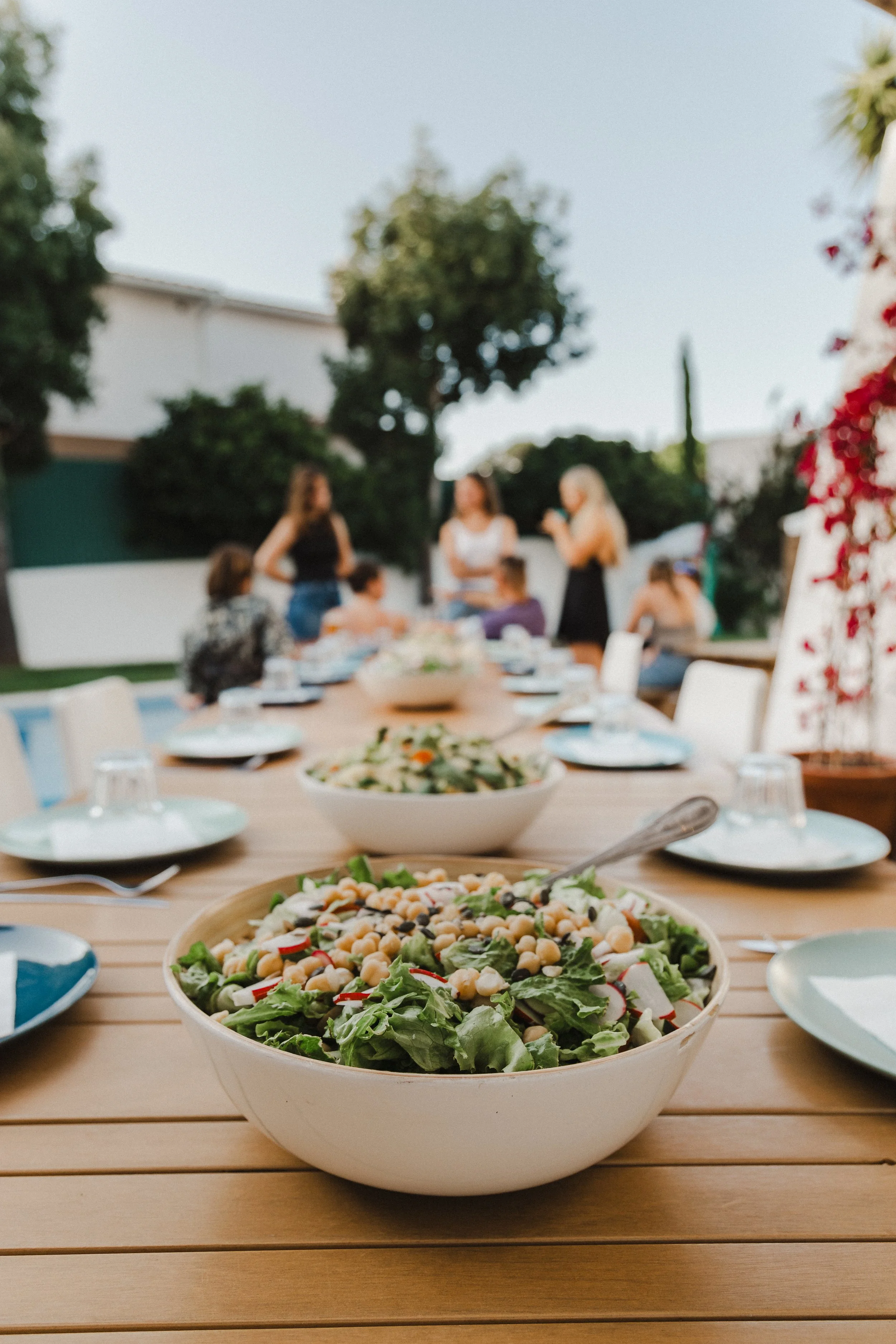 Close-up of a large salad bowl with chickpeas, greens, and vegetables on a wooden table, with other dinnerware and a blurred outdoor gathering in the background.