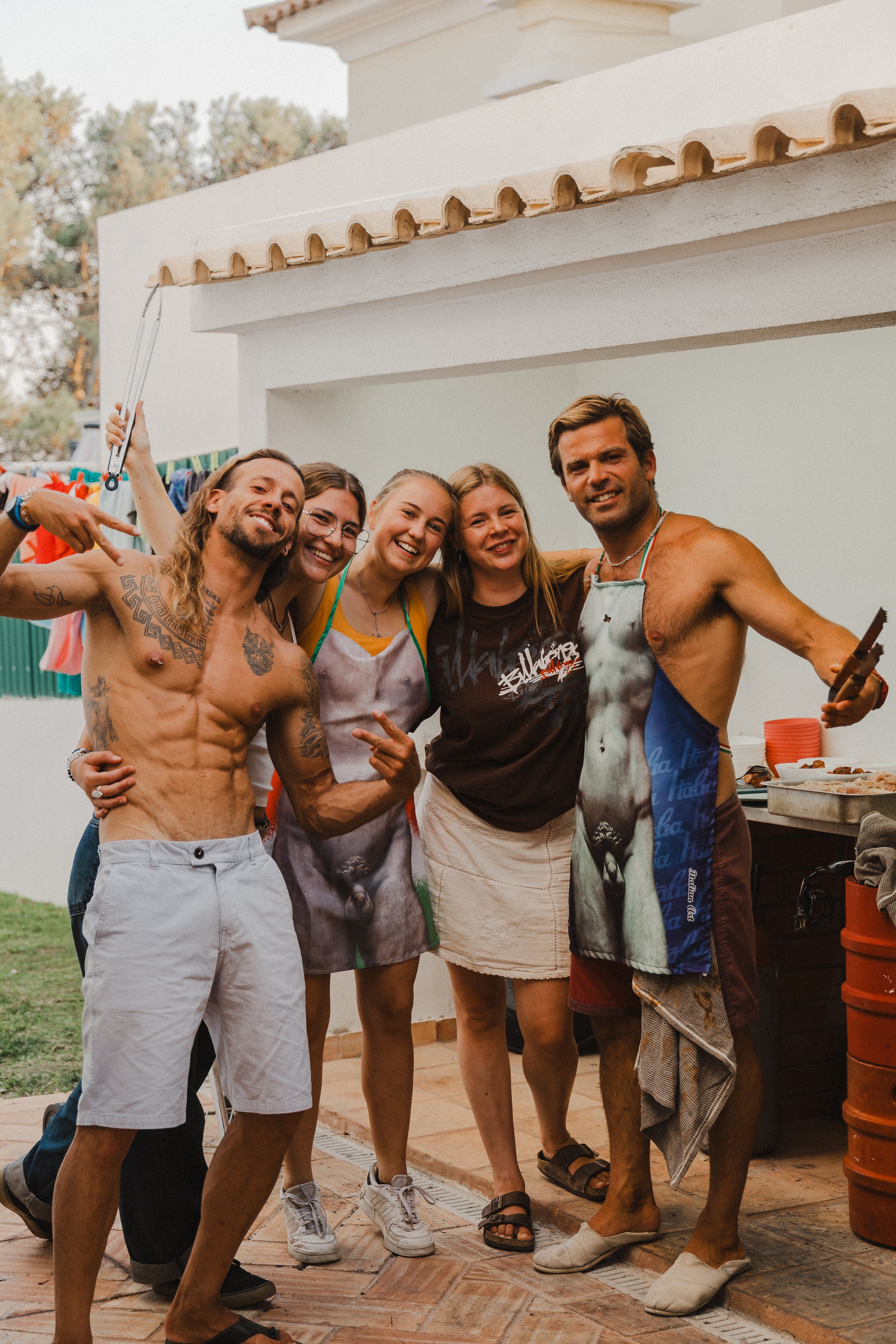 Group of six friends at a backyard barbecue, smiling and posing together. Two men are shirtless, and some people are wearing aprons. The setting is outdoors with a white building, tiled roof, and orange barrels in the background.