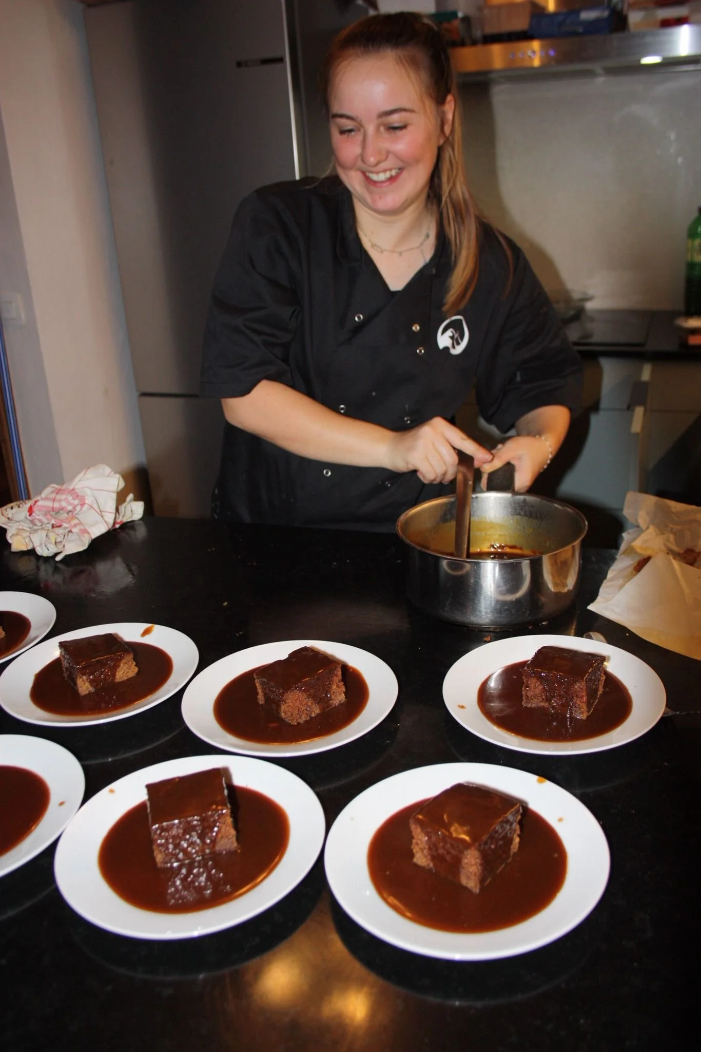 A woman in a black chef's coat preparing individual servings of chocolate cake topped with sauce at a kitchen counter.