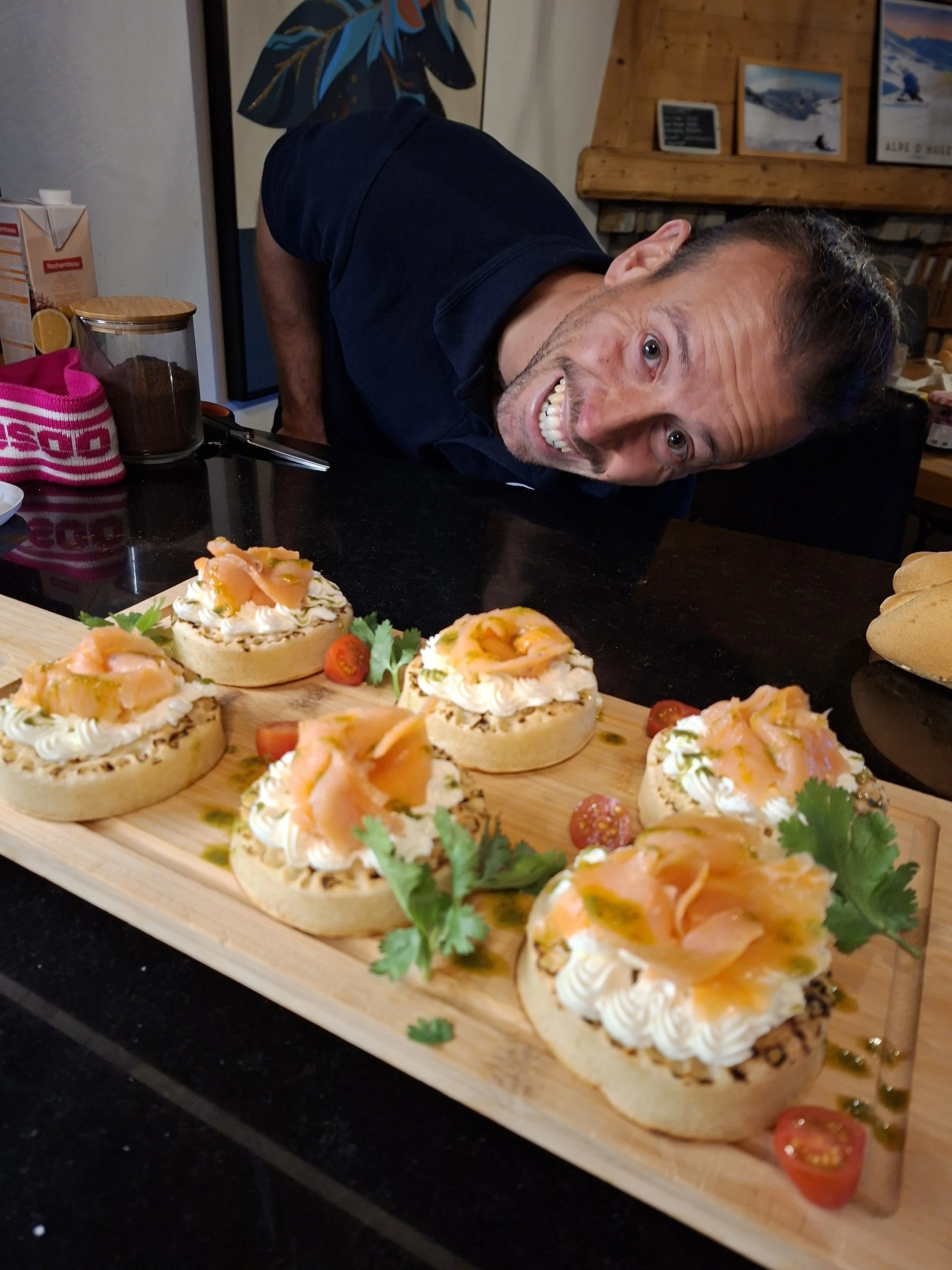 A smiling man with short hair, wearing a dark blue shirt, leaning over a wooden tray of smoked salmon canapés garnished with herbs and cherry tomatoes on a table.