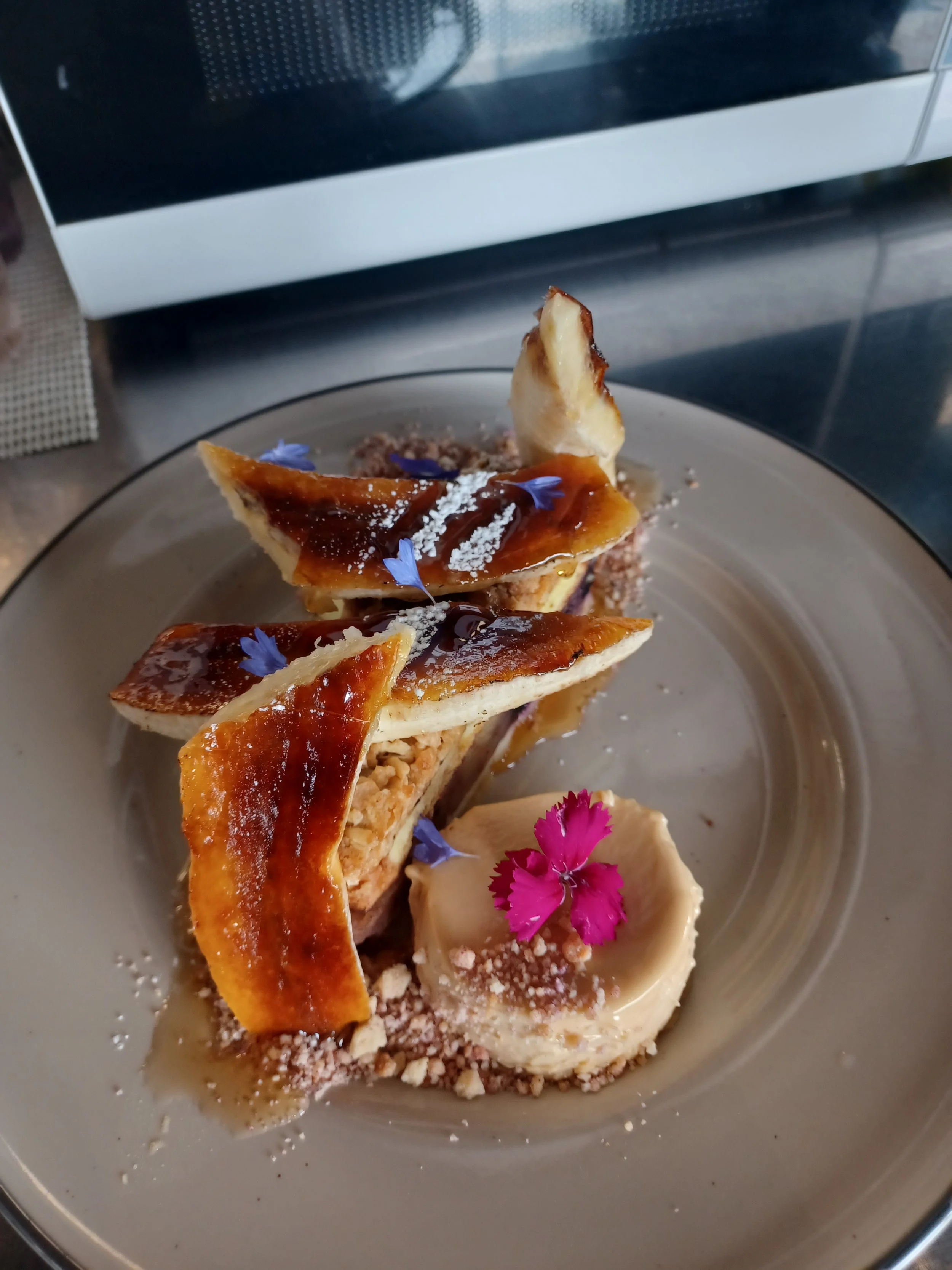A dessert consisting of a pastry topped with caramelized fruit, powdered sugar, edible flowers, and a sauce, presented on a plate decorated with edible flowers and crumbs.