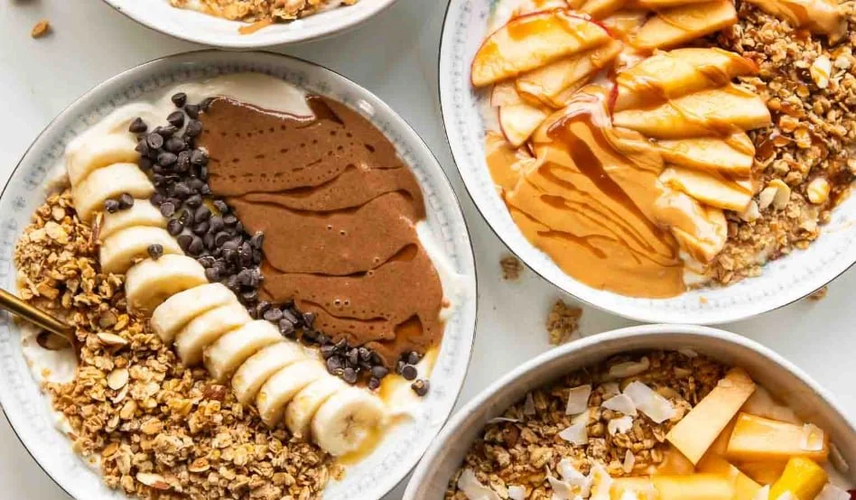 Three bowls of acai or smoothie bowls topped with bananas, granola, chocolate chips, honey, and other fruits.