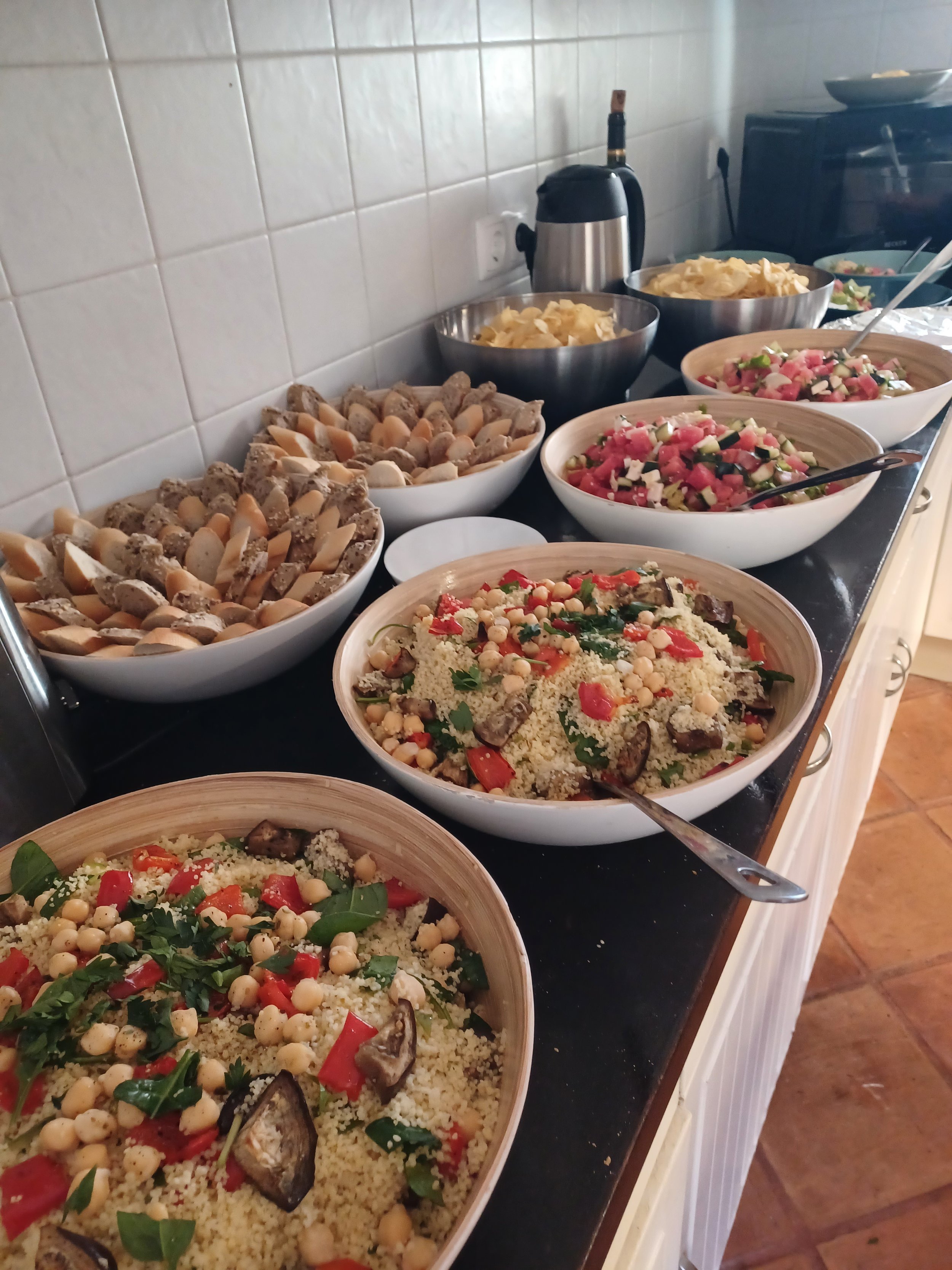 A buffet table with bowls of couscous salad, vegetable salad, potato chips, and a platter of mixed vegetables, with a kettle and a microwave in the background.