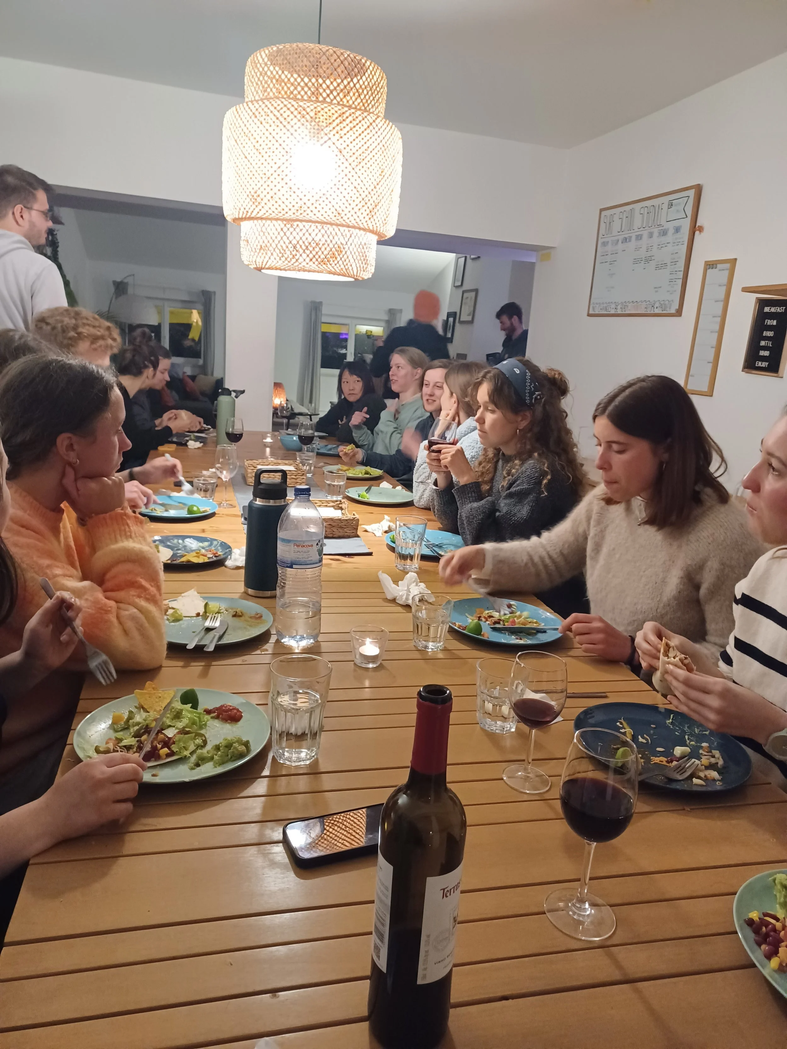 People sitting around a long dining table enjoying a meal, with plates of food, glasses of wine, and a bottle of wine in the foreground, in a cozy indoor setting with a large hanging light and framed pictures on the wall.