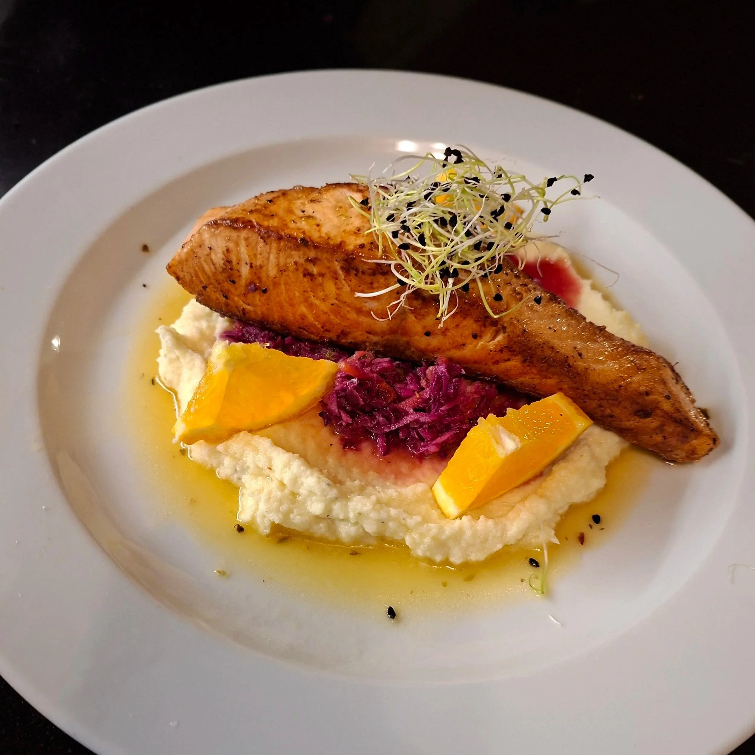 A white plate with grilled salmon topped with microgreens, served over a bed of mashed potatoes and garnished with a drizzle of yellow sauce and red beet chutney.