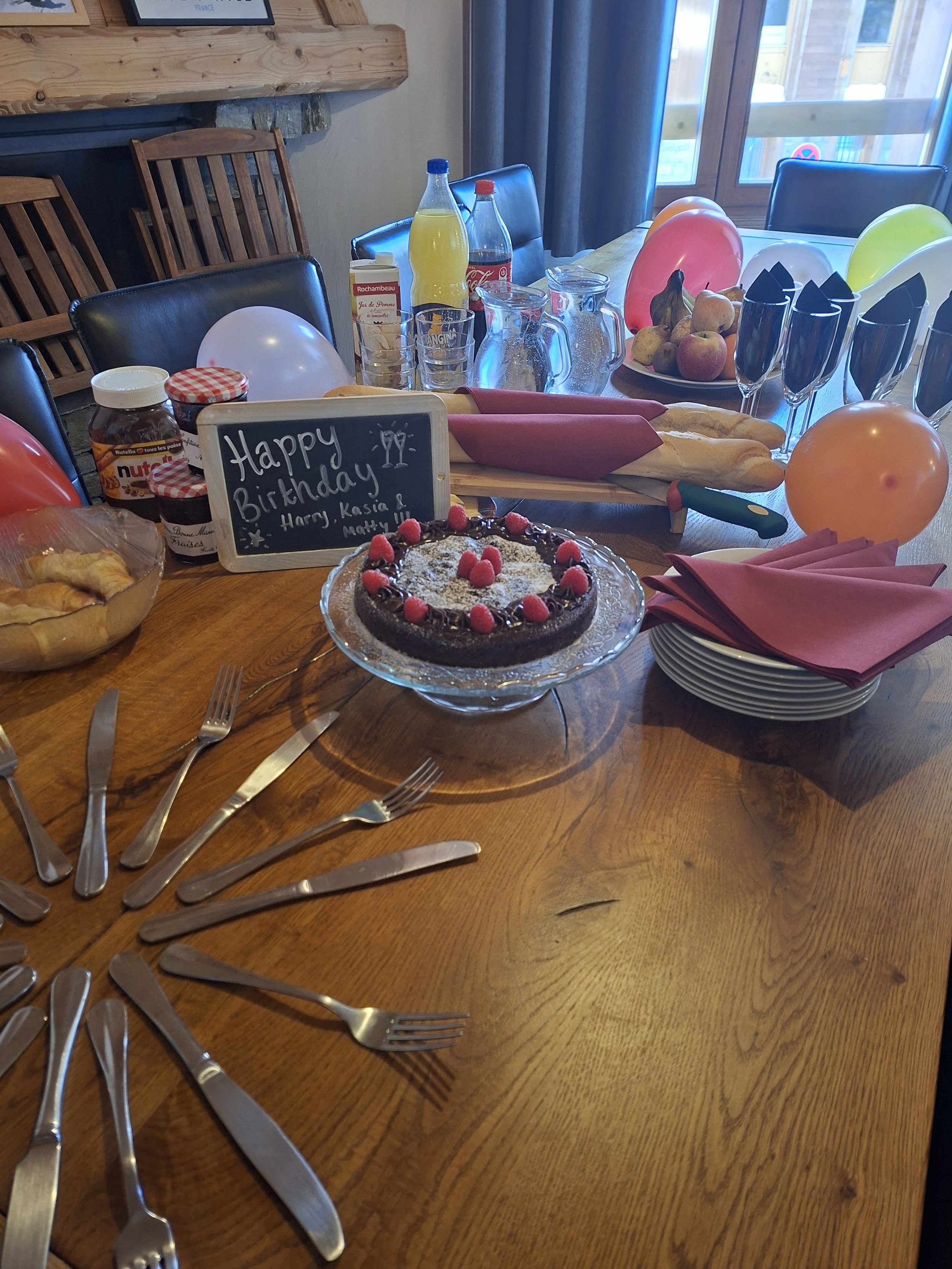 A decorated table set for a birthday celebration with balloons, a cake, food, drinks, utensils, and a chalkboard sign reading 'Happy Birthday Harry, Kasia & Matty' inside a wooden cabin.