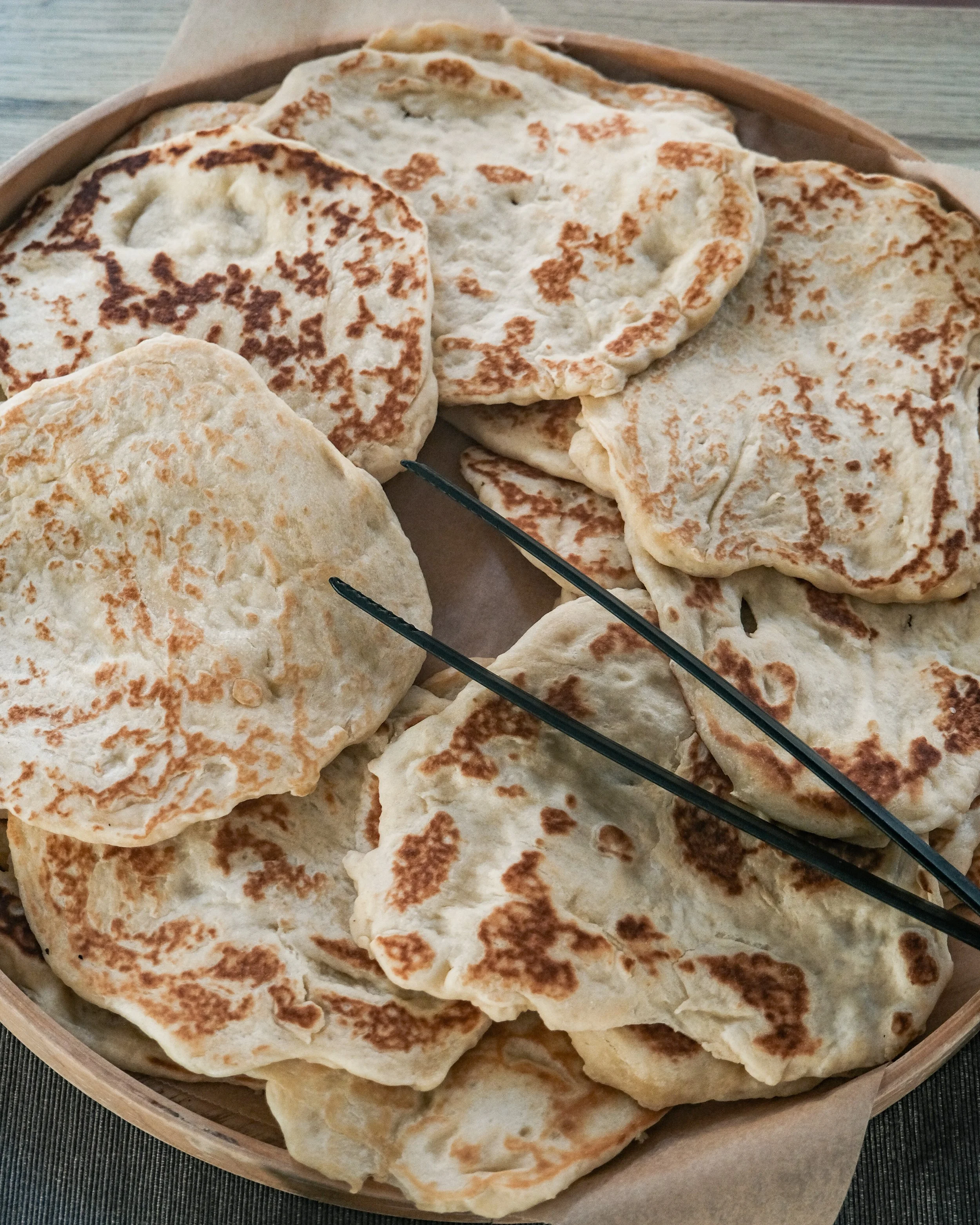A basket of freshly cooked flatbreads, likely naan or similar type, lined with parchment paper.