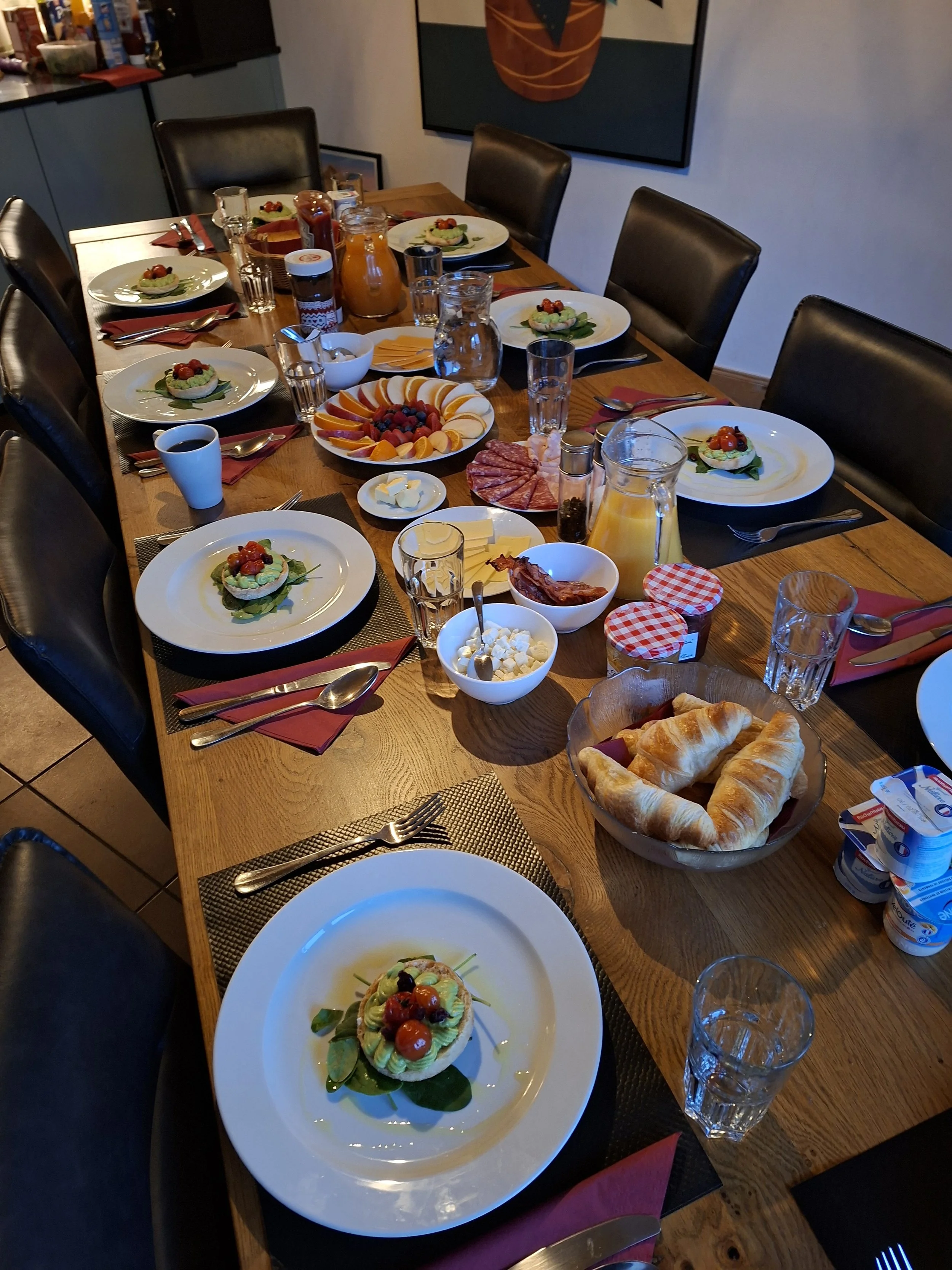 A dining table set for a meal with plates of food, glasses of water, utensils, and bowls of condiments, with a plate of croissants at the front of the table.