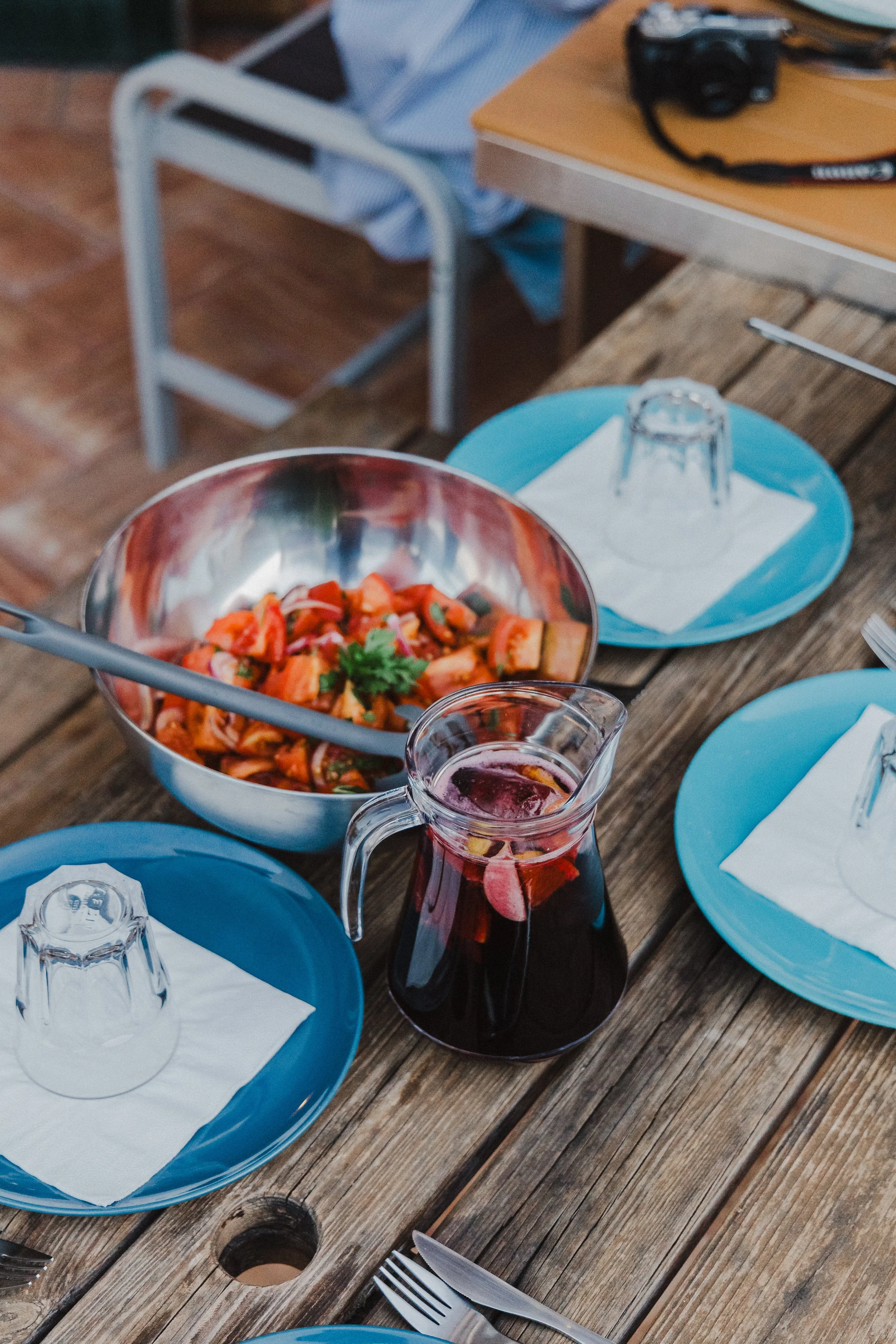 A wooden outdoor table set with blue plates, clear glasses, a pitcher of dark red beverage, and a metal bowl of salad with vegetables and herbs. There are also white napkins and cutlery on the table.