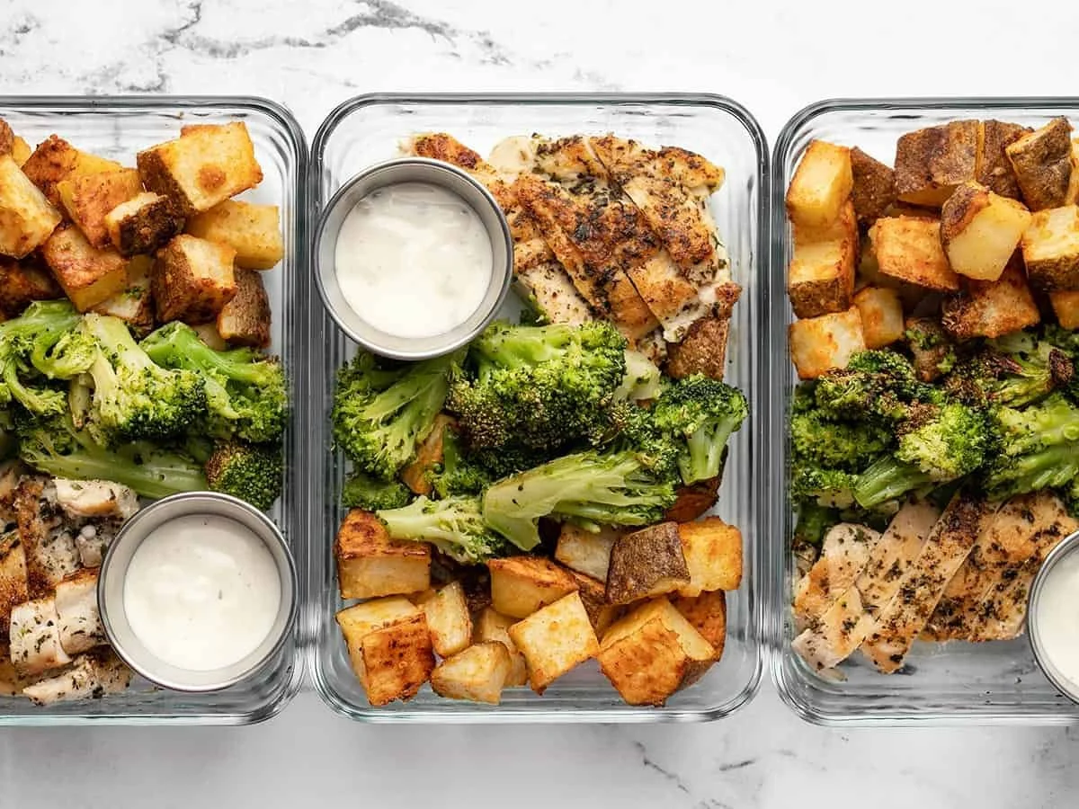 Three glass meal prep containers filled with roasted broccoli, grilled chicken, roasted potatoes, and a side of creamy dressing, placed on a white marble surface.