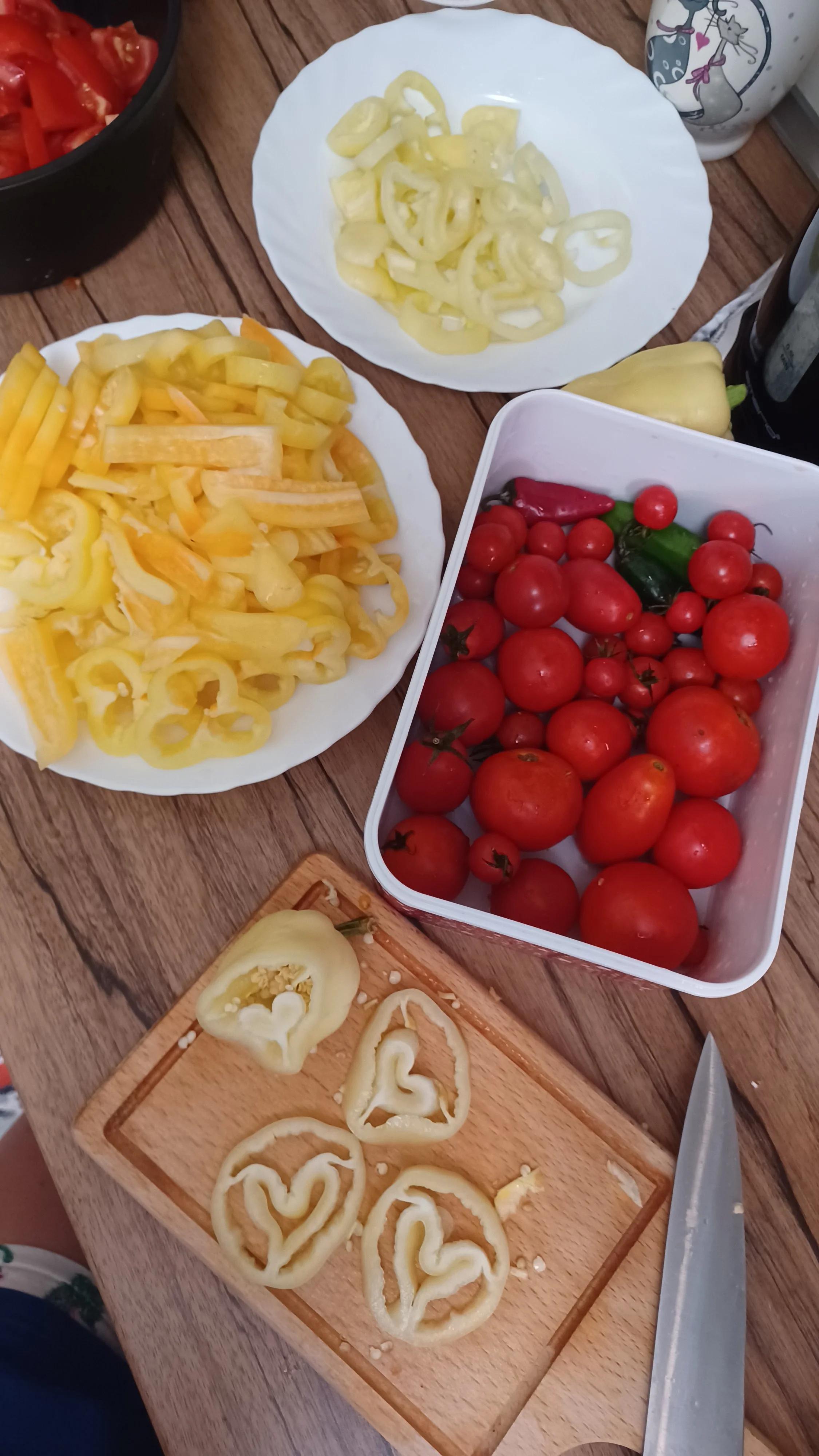 Assorted fresh vegetables on a wooden table including sliced bell peppers, tomatoes, and yellow onions.
