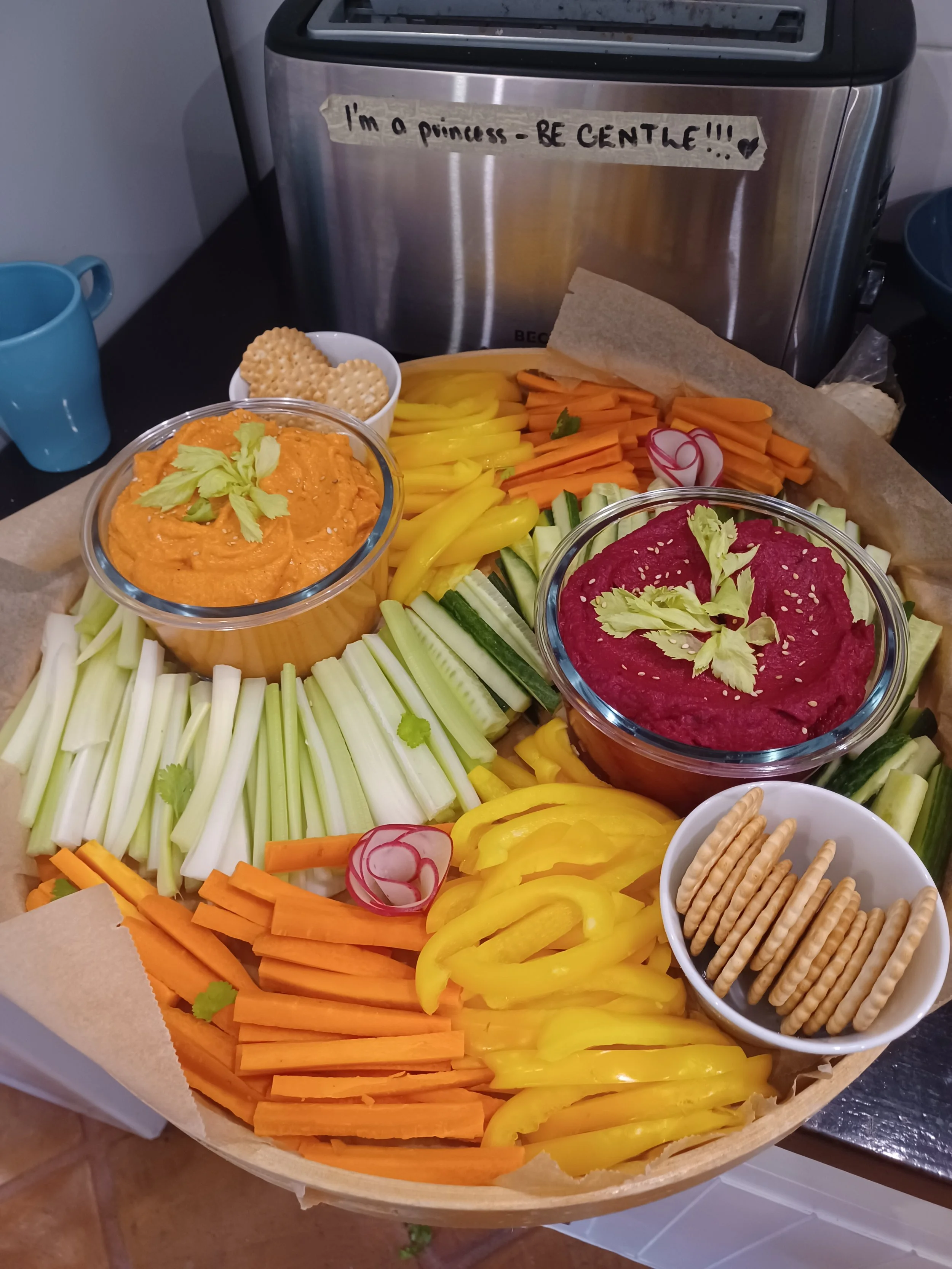 A wooden tray filled with sliced vegetables including celery, carrots, yellow bell peppers, cucumber, and radishes, with bowls of hummus, beet hummus, and crackers on top.