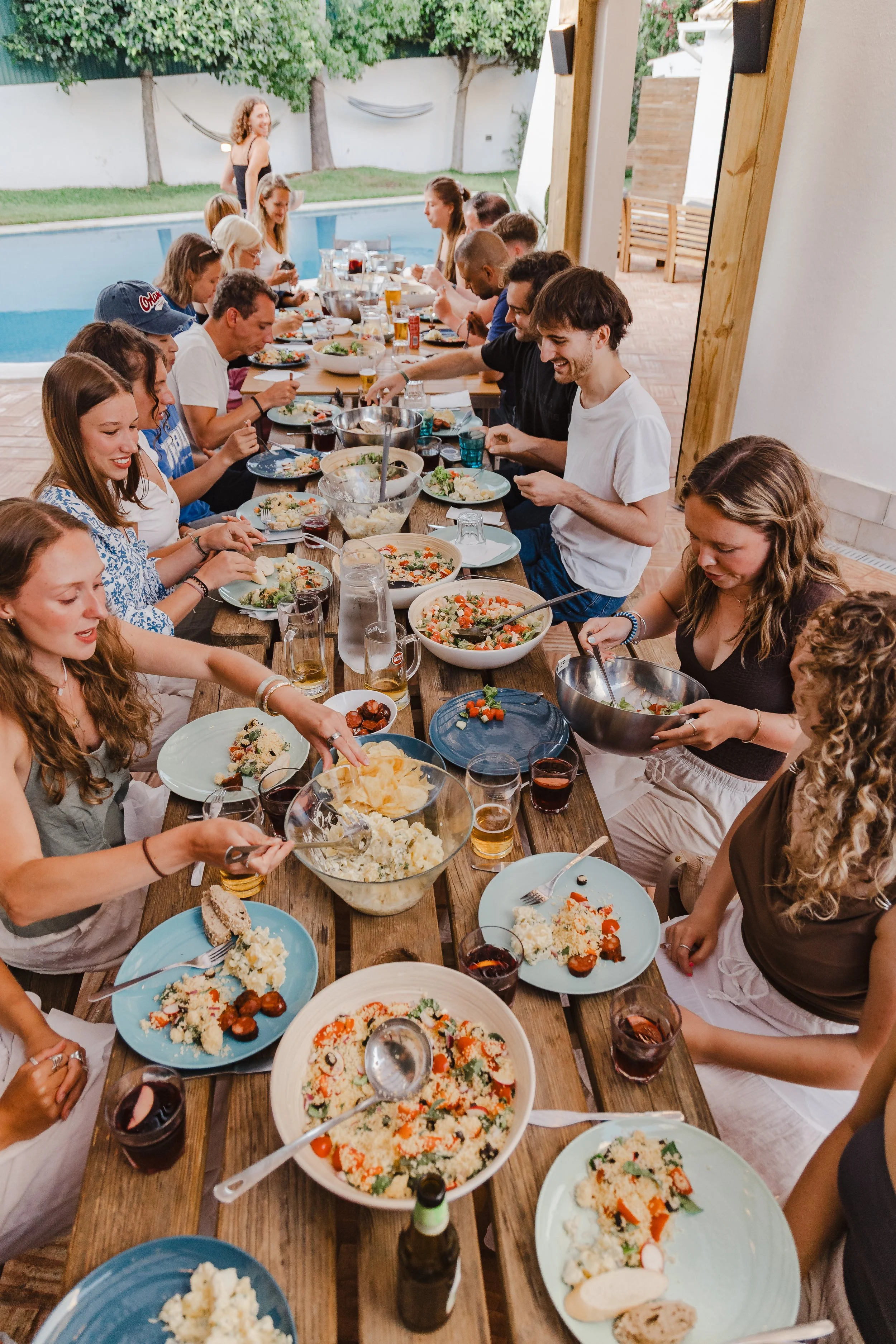 People enjoying a meal at a long wooden table outdoors near a pool, with dishes like salads, rice, and drinks, on a patio with trees and a hammock in the background.
