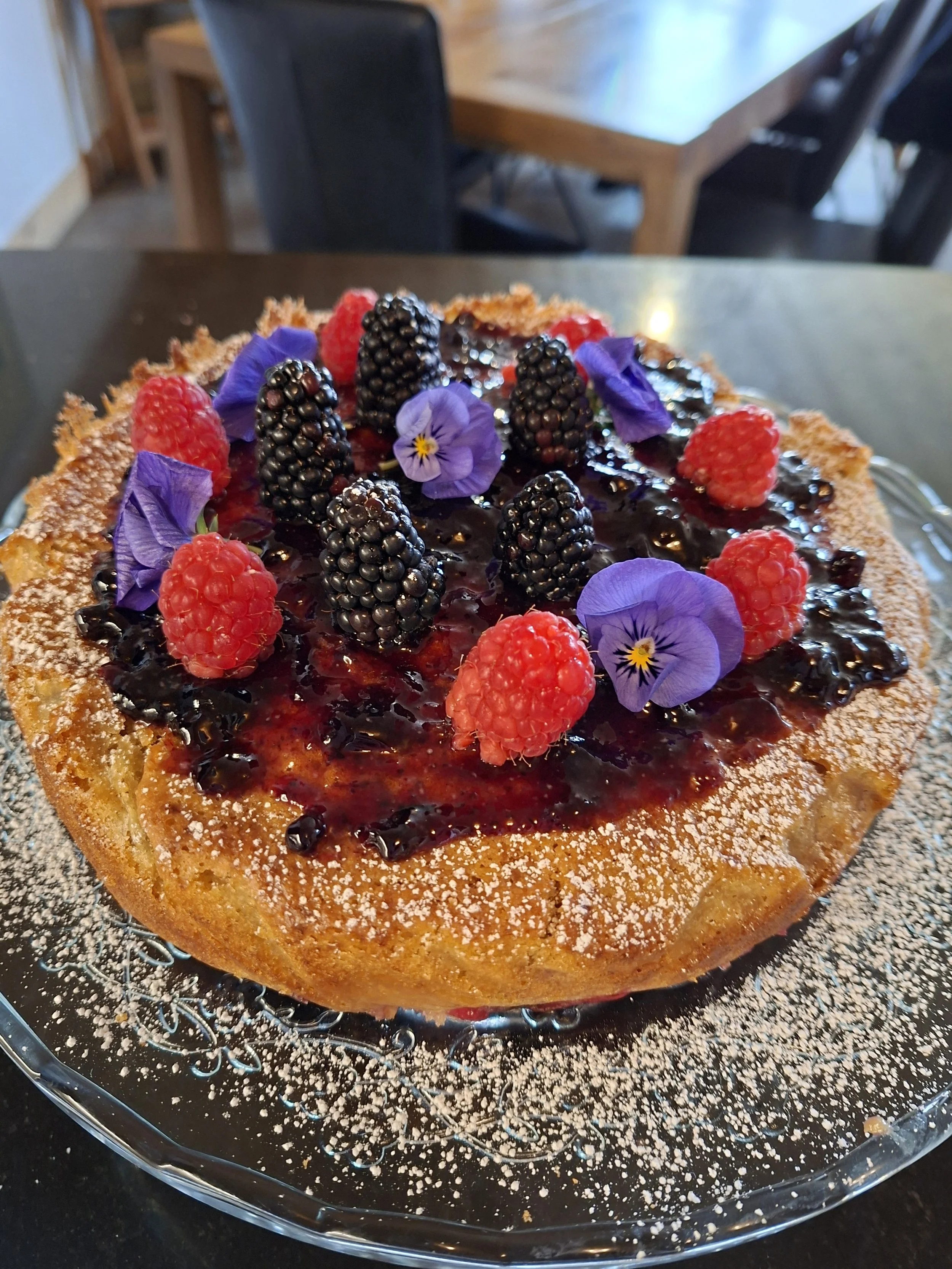 Close-up of a cake topped with blackberries, raspberries, violet edible flowers, and a shiny glaze, on a glass plate with powdered sugar around the edge.