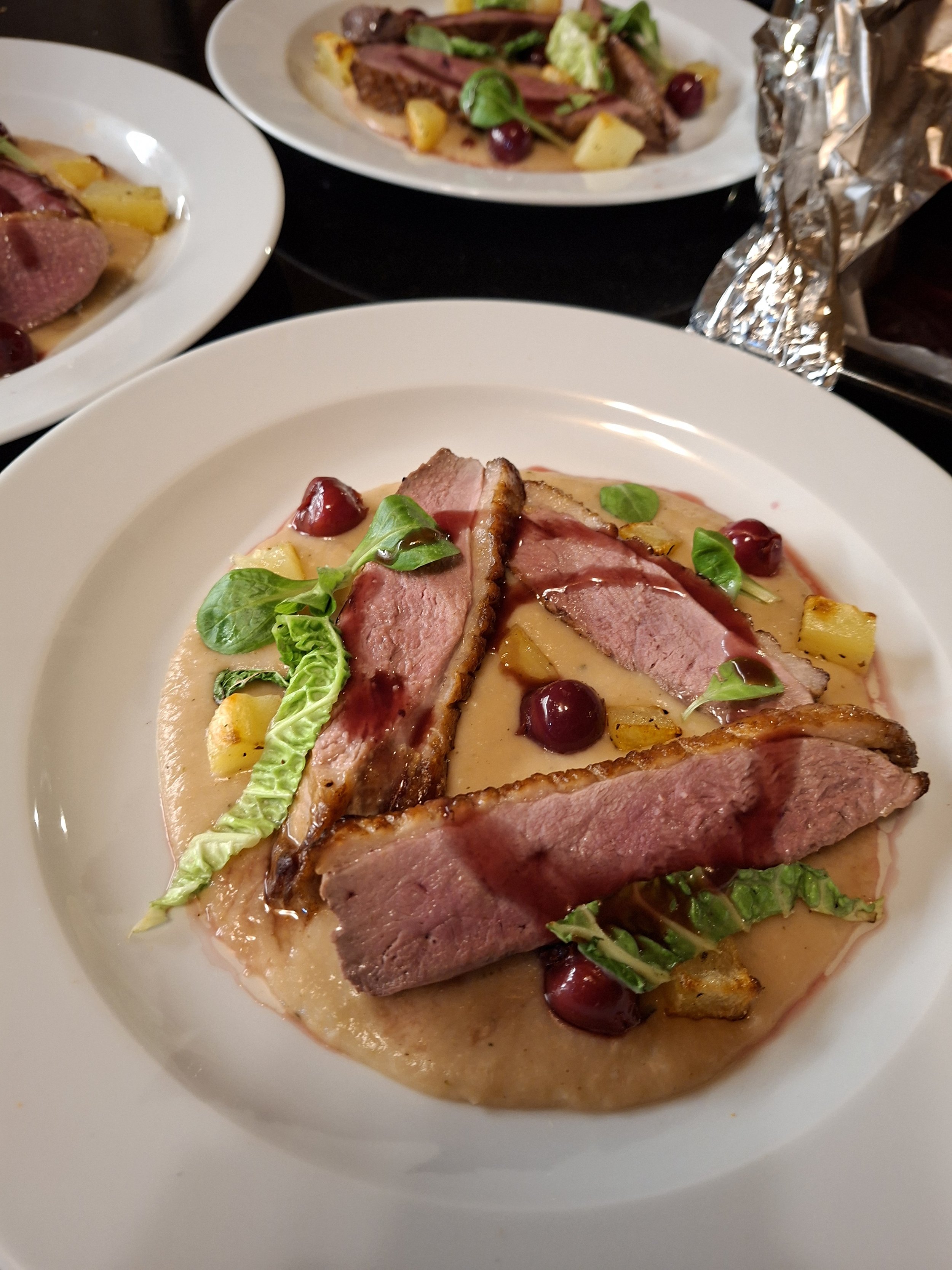 A white plate with medium-rare slices of beef, garnished with green leaves, surrounded by a creamy sauce with chunks of pineapple, red berries, and herbs.