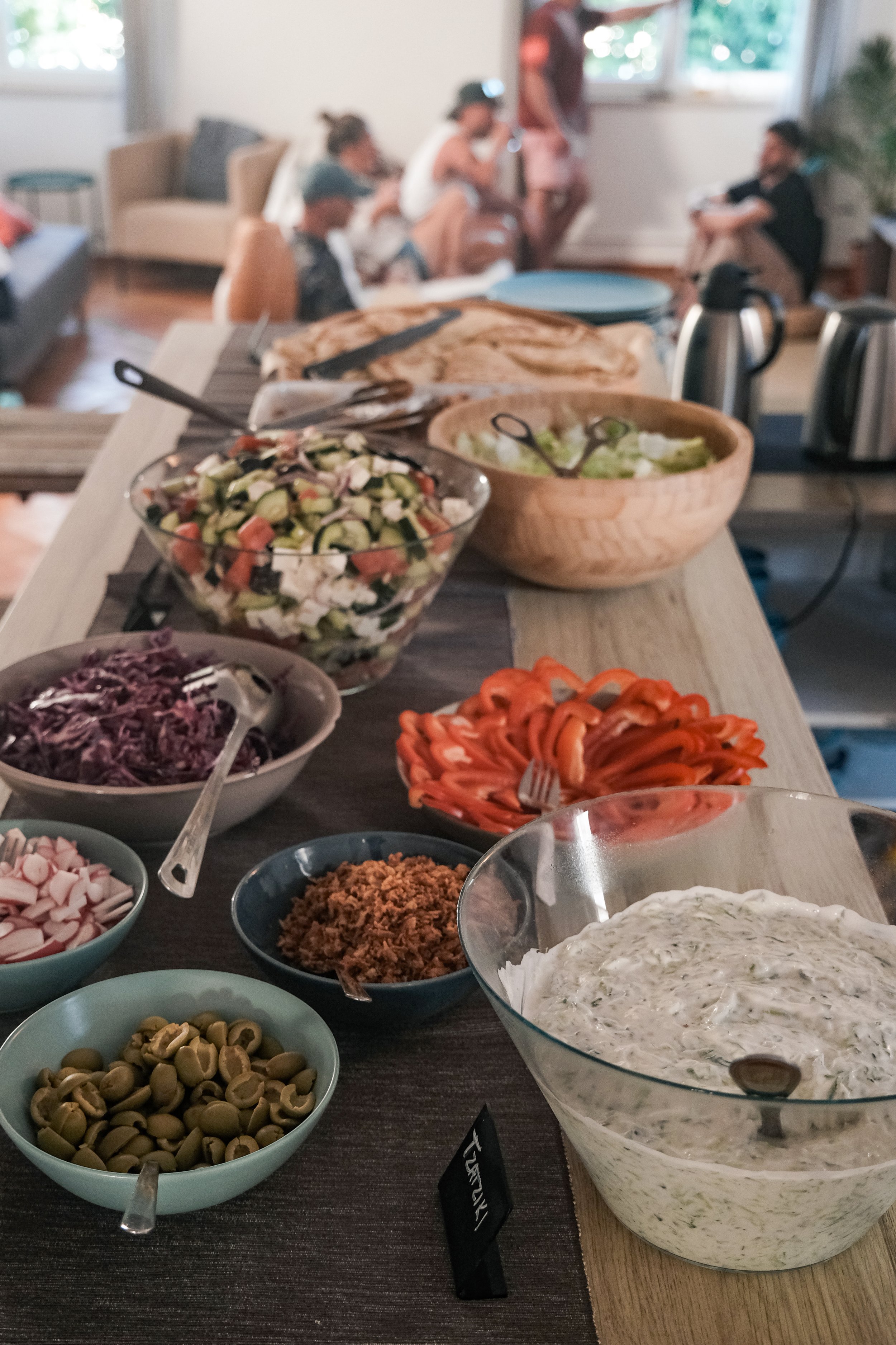 Assorted bowls of salad, pickles, olives, shredded vegetables, and dips on a table in front of a blurry background of people sitting on couches and chairs in a living room.