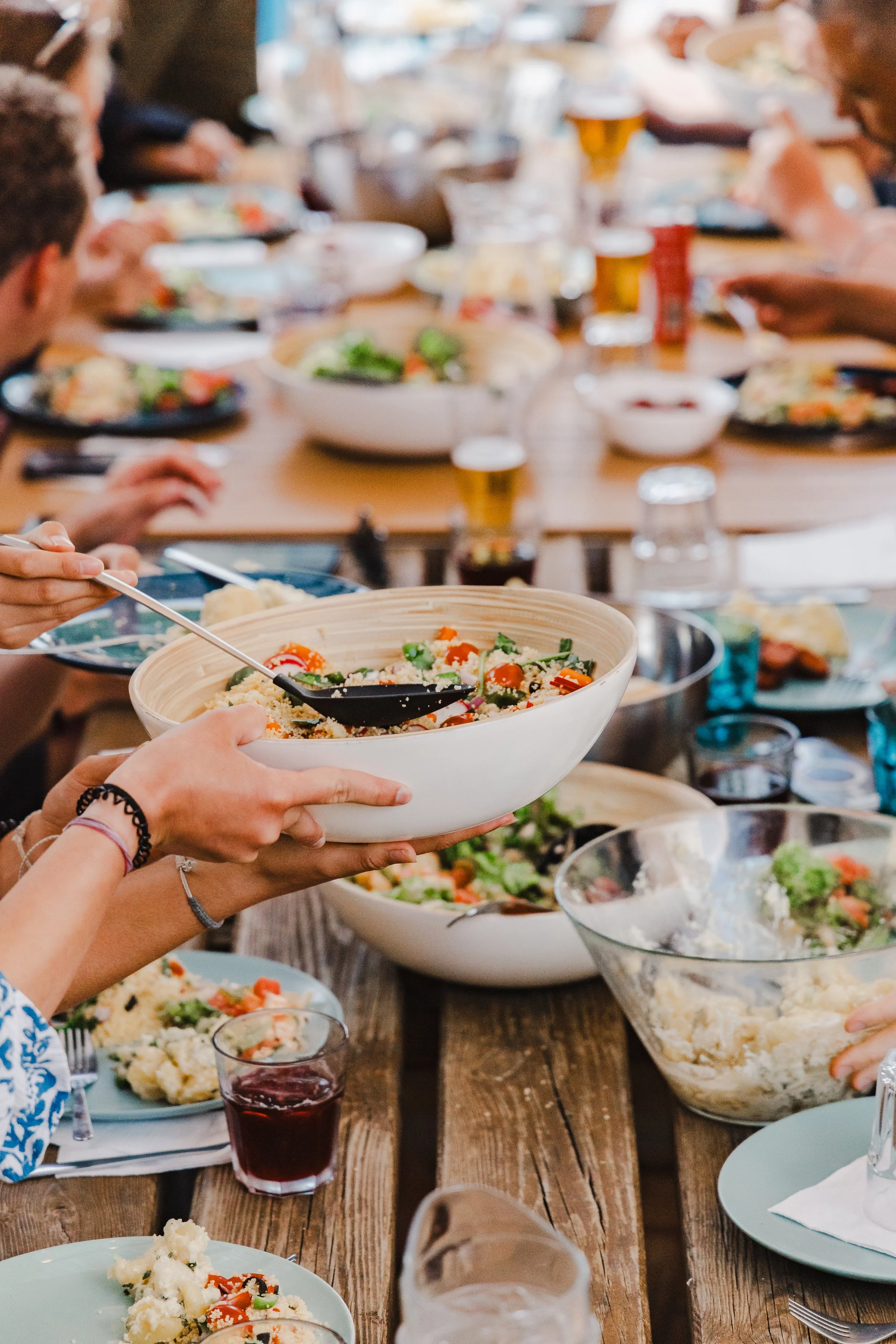 People gathered around a wooden table enjoying a meal with various dishes, salads, and glasses of beverages at a social gathering.