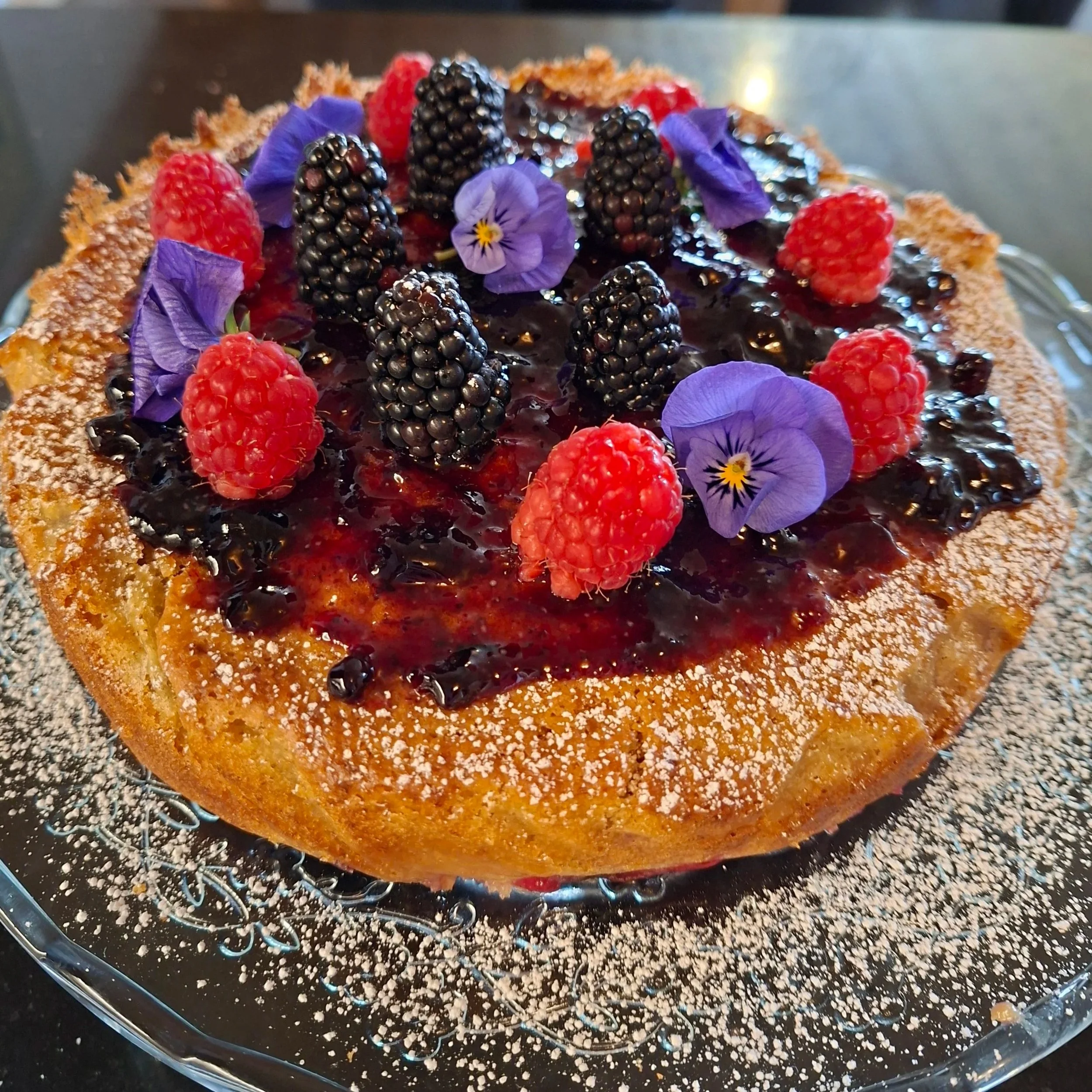 A close-up of a dessert topped with blackberries, raspberries, and purple edible flowers, with a crusty edge and glossy surface.