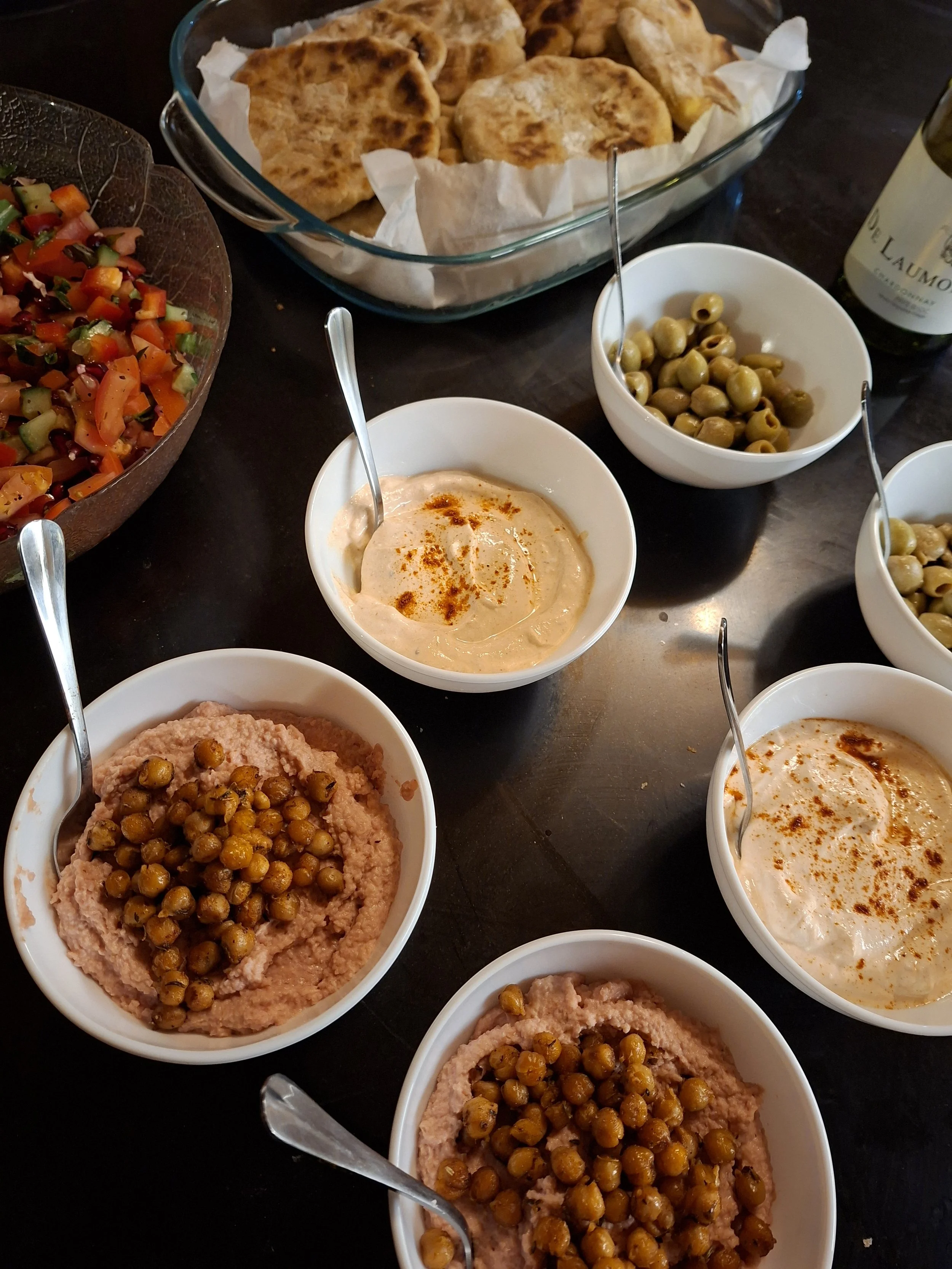 A table with several bowls of hummus topped with sumac and whole chickpeas, a large glass dish of flatbreads, small bowls of green olives and creamy dip, a salad with chopped vegetables, and a bottle of wine.